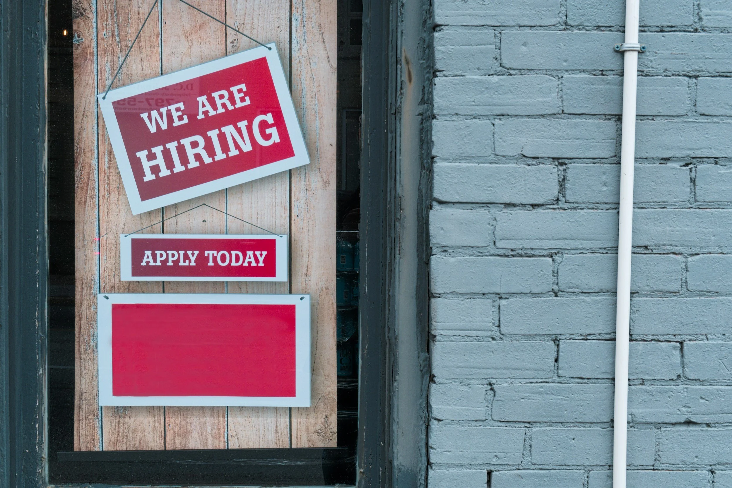 Sign on a window advertising job openings, with text 'We Are Hiring', 'Apply Today', and a blank space for additional information, set against a brick wall background.