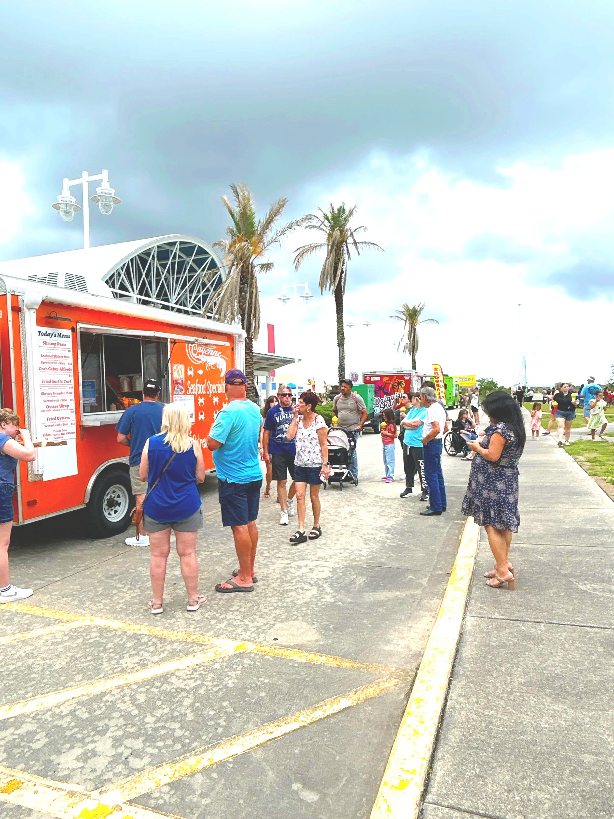 People waiting in line at food trucks in an outdoor area with palm trees and cloudy sky.