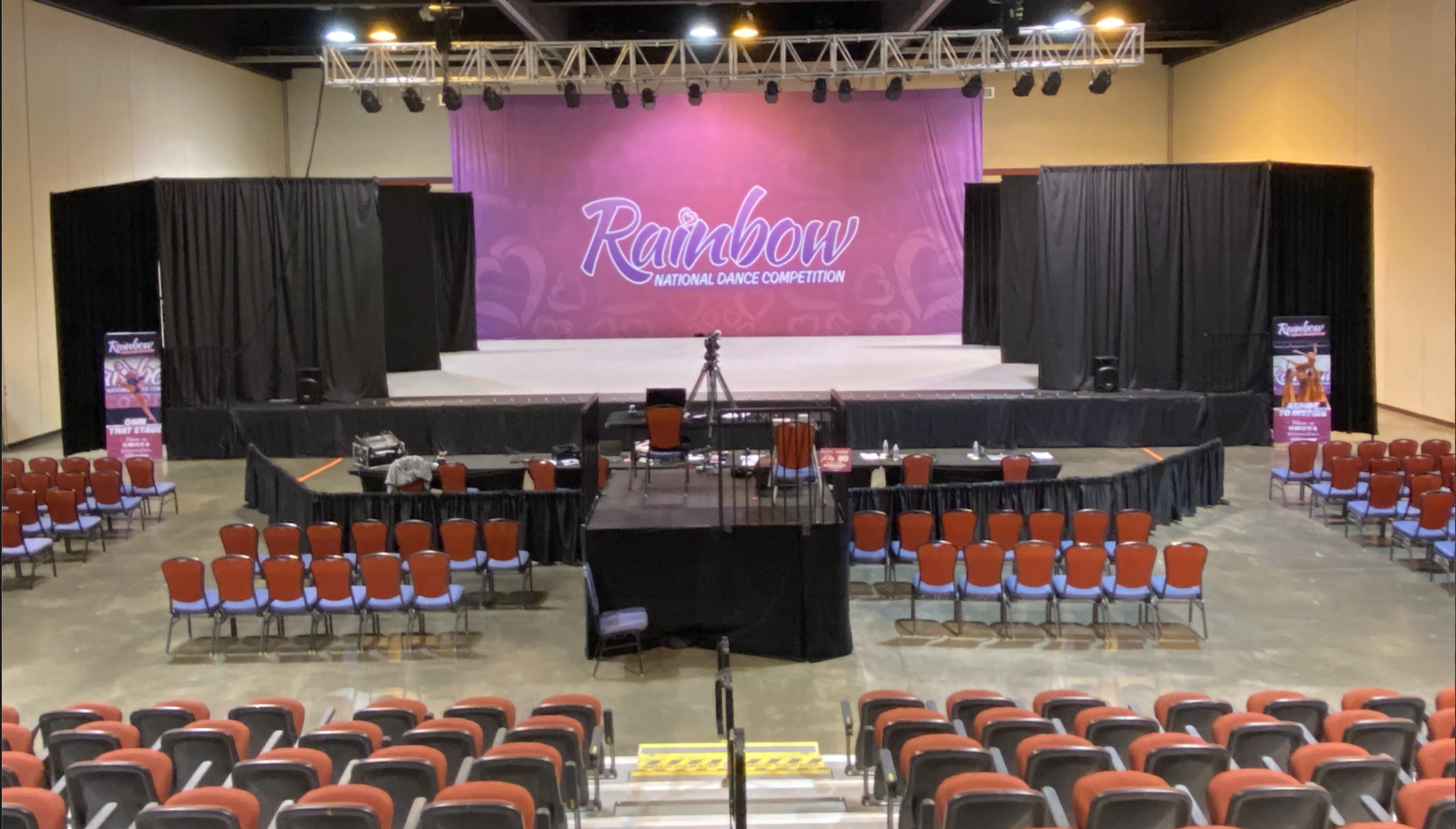 Empty stage at Rainbow National Dance Competition with a large pink backdrop, black curtains on either side, rows of red and black chairs in front, and a camera on a tripod set up in the center.
