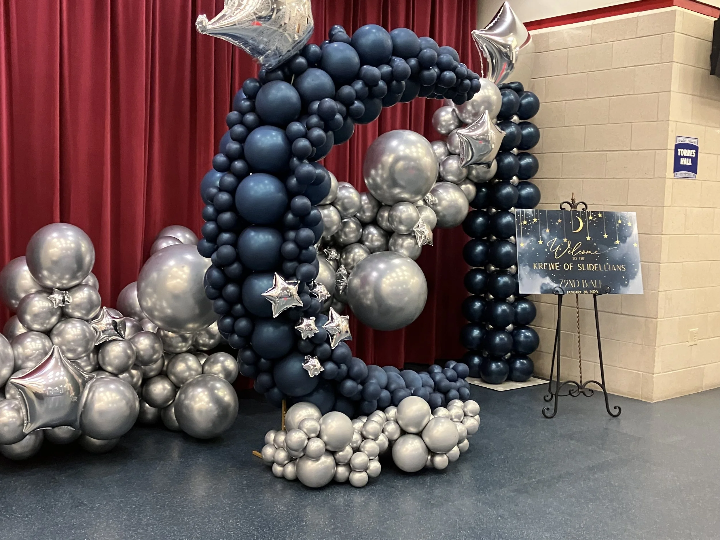 Balloon arch with blue, silver, and star-shaped balloons at an event entrance, with a welcome sign on an easel nearby.