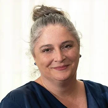 A woman with gray hair in a bun, wearing a navy blue top, smiling gently, standing in front of a light-colored background.