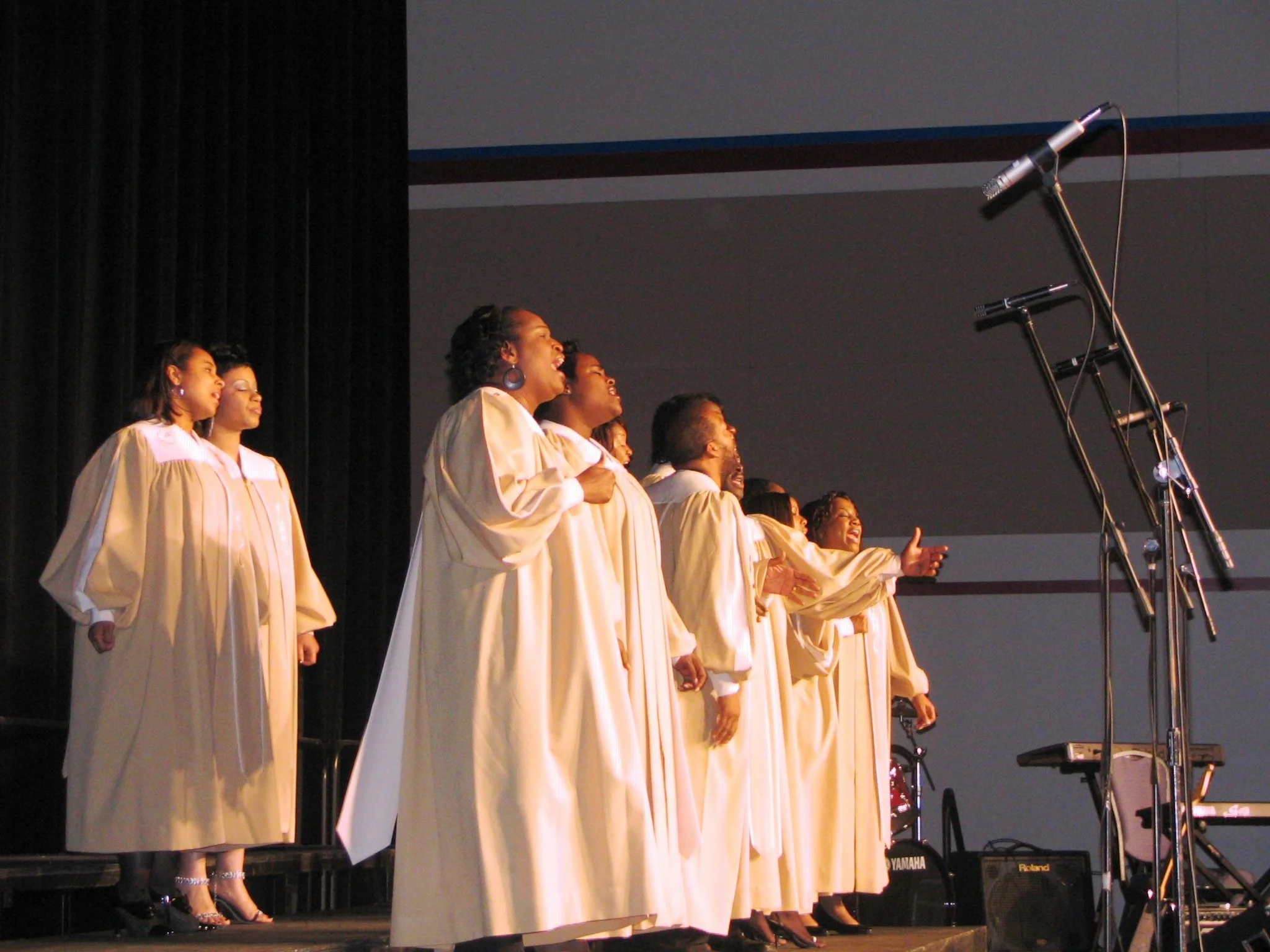 Church choir dressed in beige robes singing on stage with microphones.