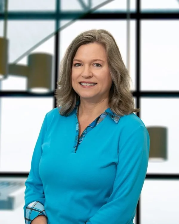 A woman with shoulder-length gray hair smiling stands in front of a large window in an indoor setting.