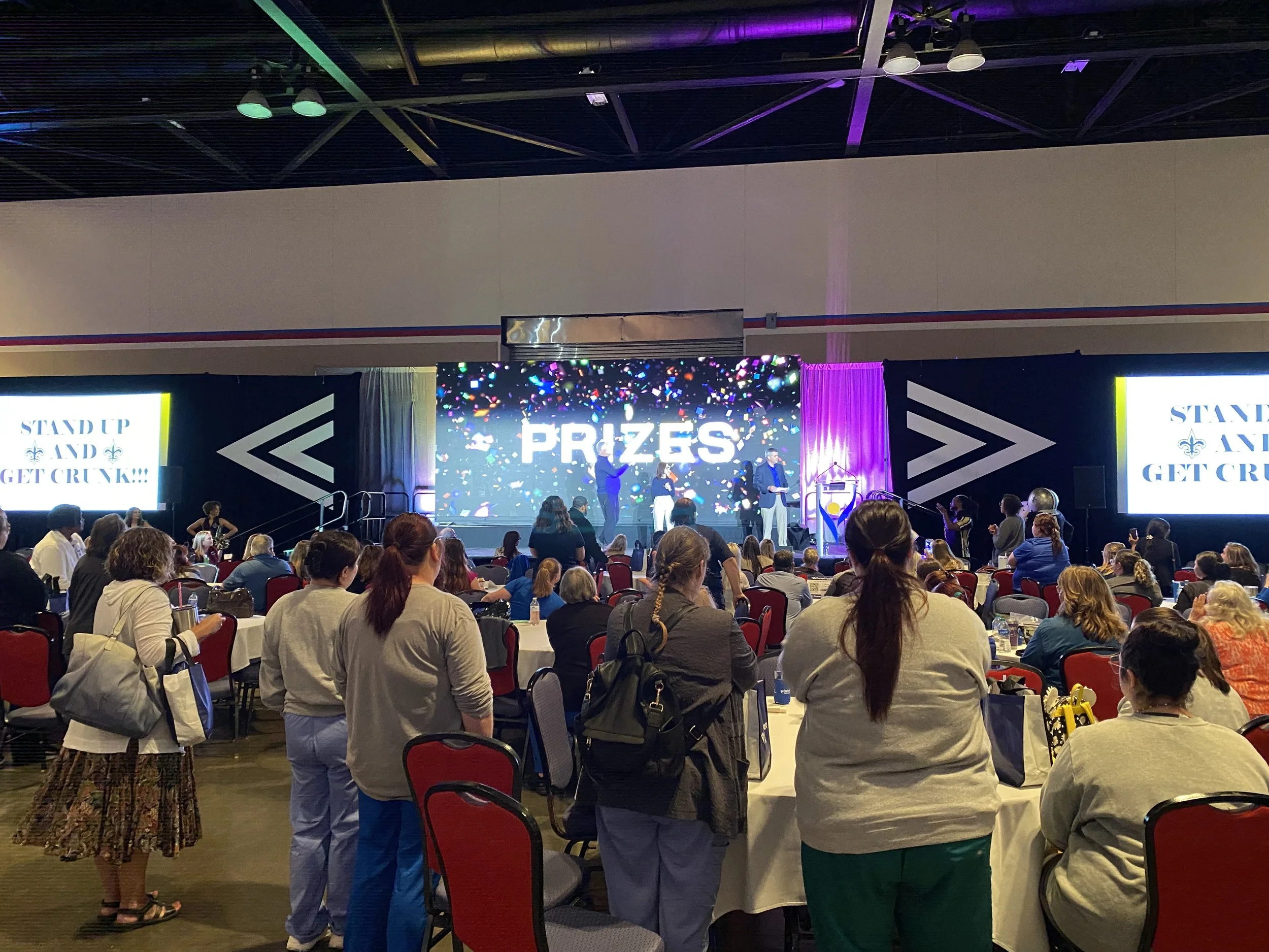 A large indoor event with a stage displaying the word 'PRIZES' and screens showing messages like 'STAND UP AND GET CRUNK!!!'. Venue filled with round tables and seated audience, some standing, with people watching a presentation or award ceremony.