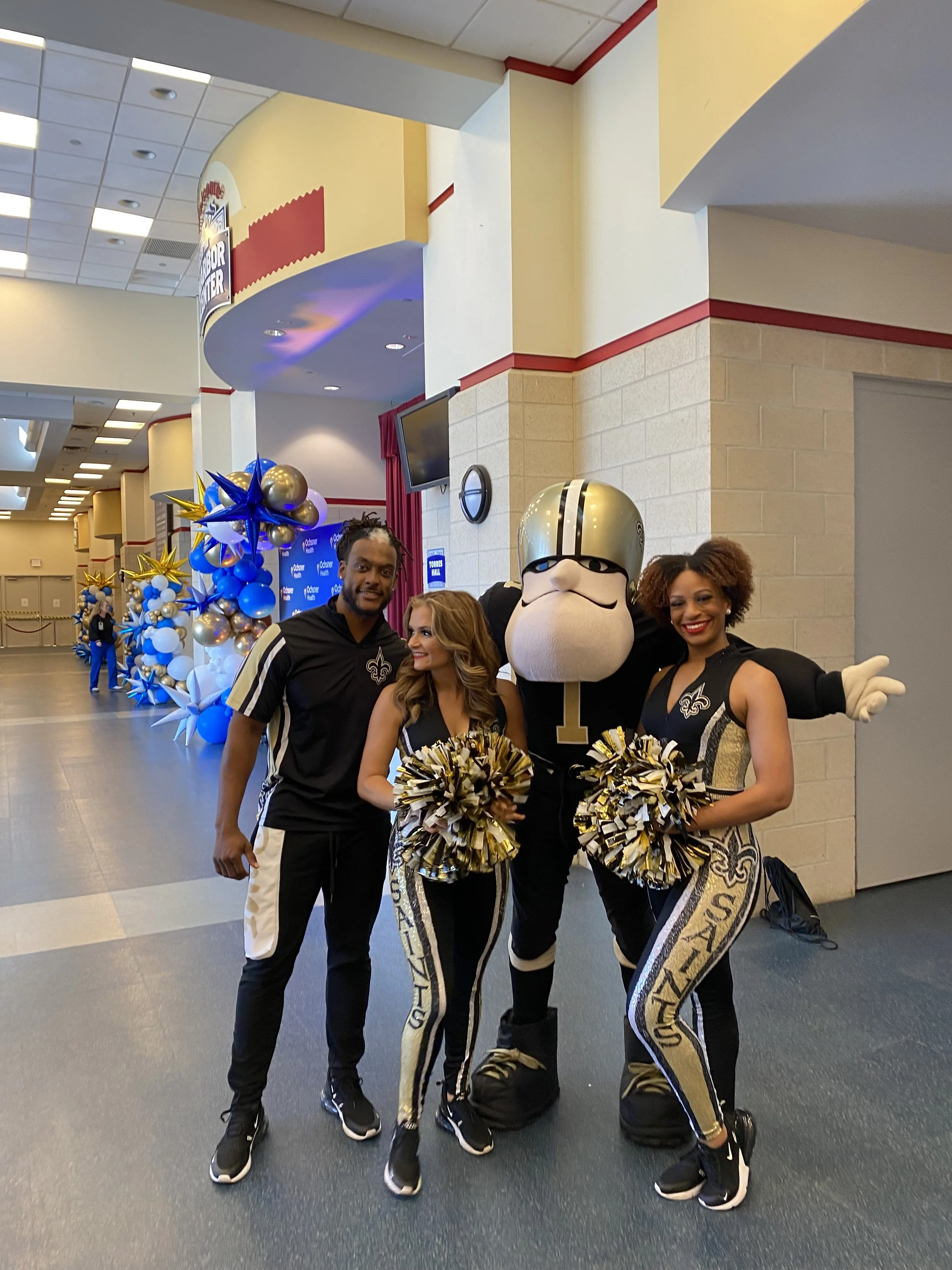 Group of four people at a sports event, including a mascot and cheerleaders in New Orleans Saints attire, with decorations and balloons in the background.