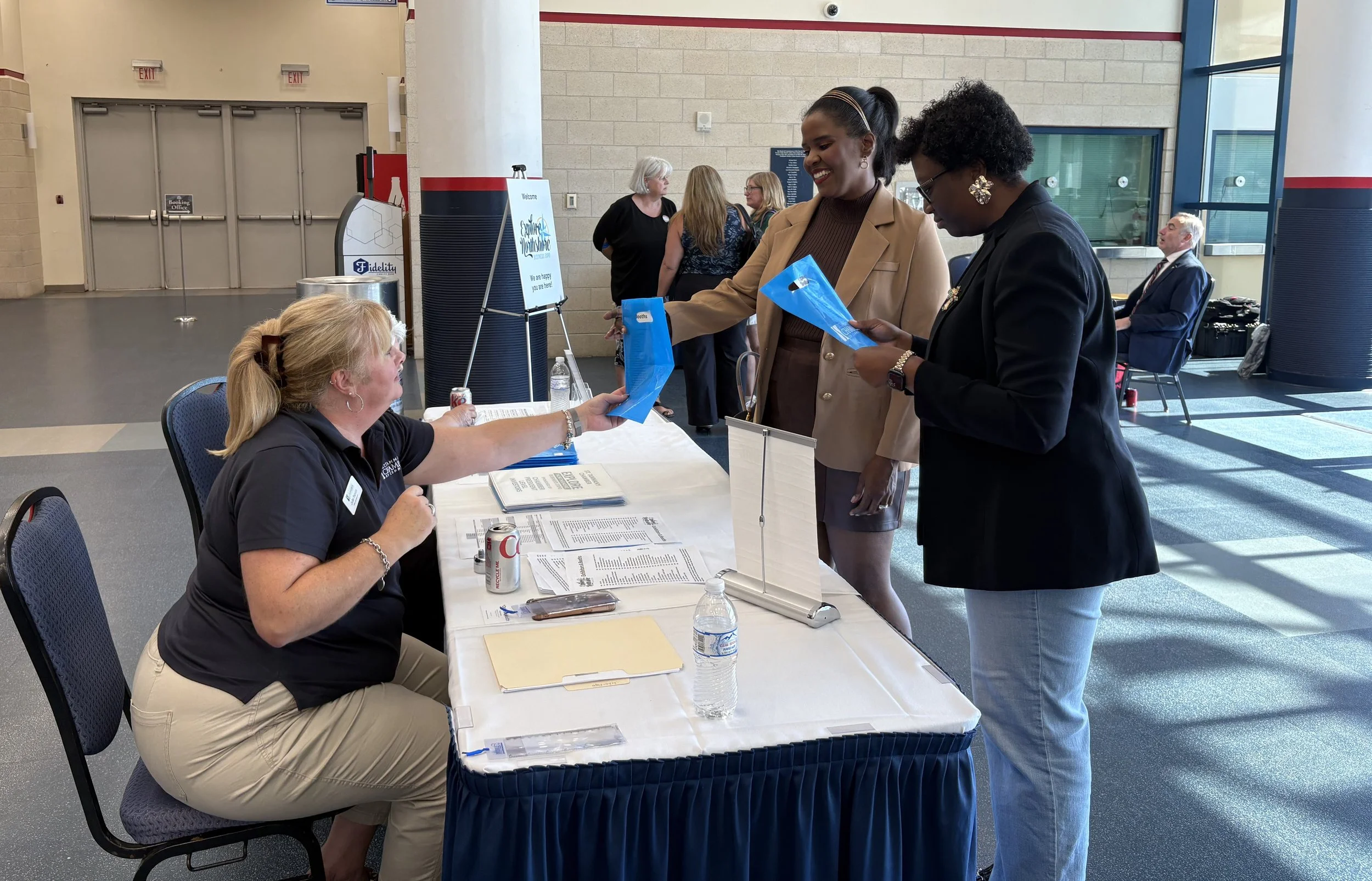 Two women are at a registration table; one woman is handing an item to the other woman who is seated. Several other people are in the background inside a large, well-lit building, conference or event center, with a registration sign and informational