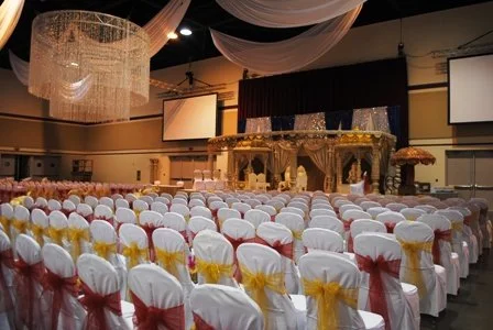 Indoor event venue set up for a ceremony with rows of white chairs decorated with pink and yellow sashes, a stage with golden ornamental arch, draped fabric ceiling, and large chandeliers.
