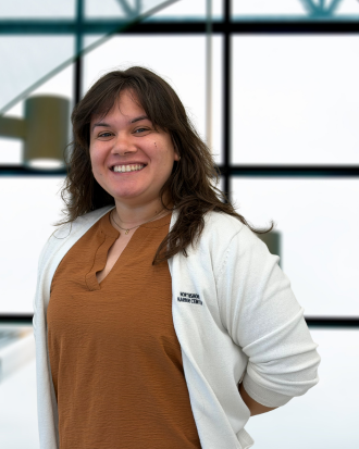 Smiling woman with dark hair in a white coat standing in front of a modern office background.