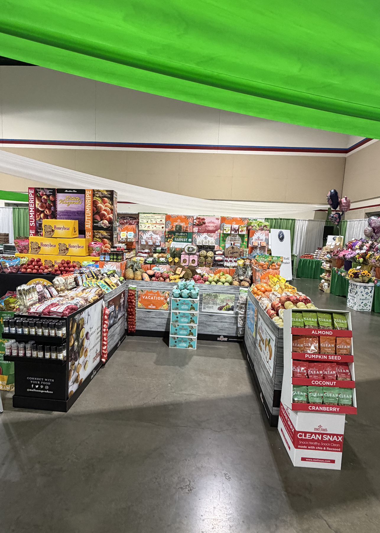 Display of various fruits and packaged snacks at an indoor market or fair, with banners and signs.