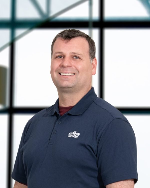 Smiling man in navy polo shirt with 'Harbor Center' logo, standing in front of large window with grid framing in an modern indoor setting.