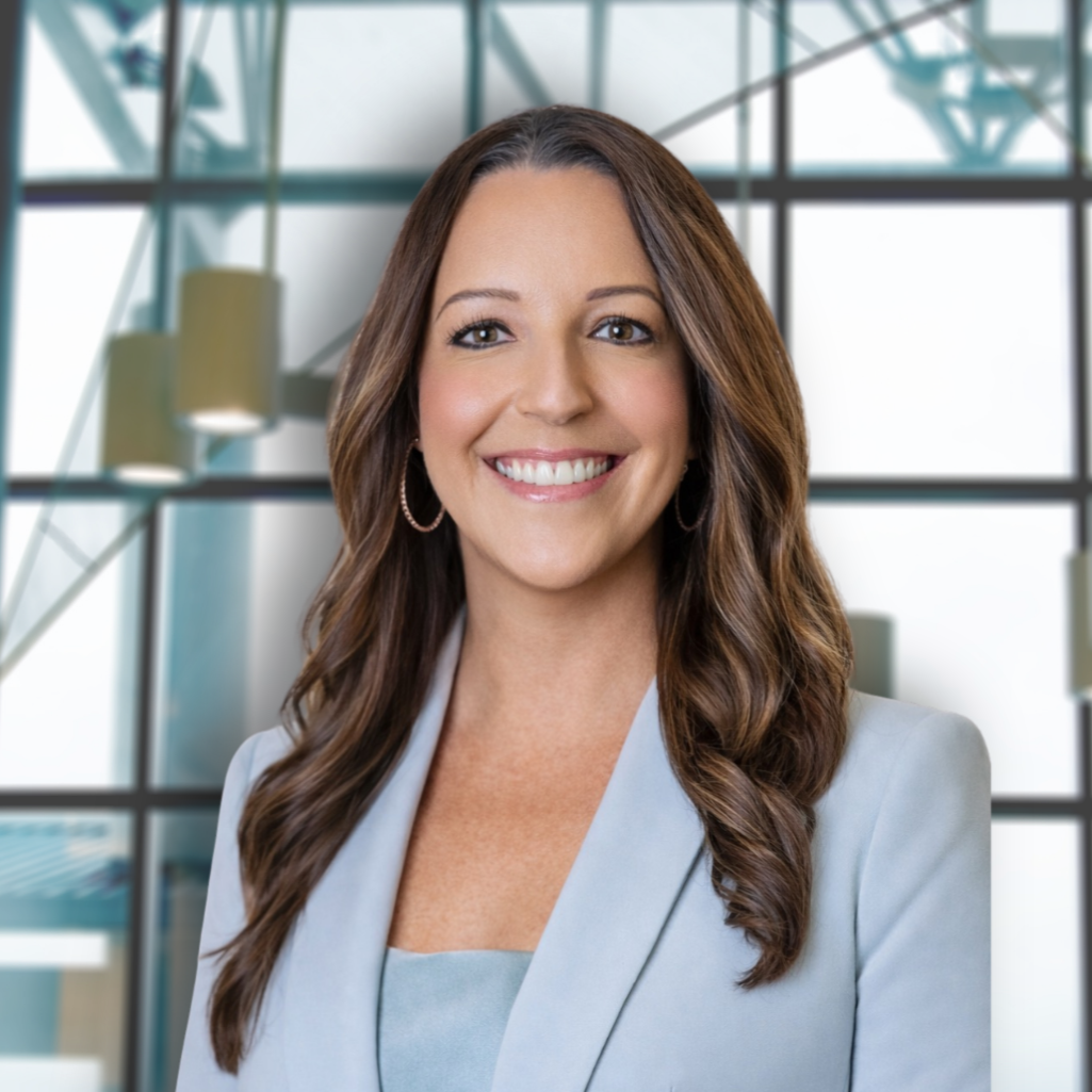 A smiling woman with long brown hair in a light grey blazer, standing in front of a large glass window in an office building.