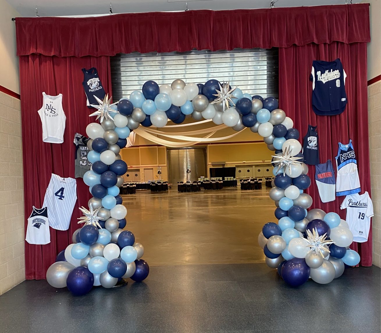 Decorated archway with blue, white, and silver balloons, hockey jerseys on curtain, and a sports-themed event hall in background.