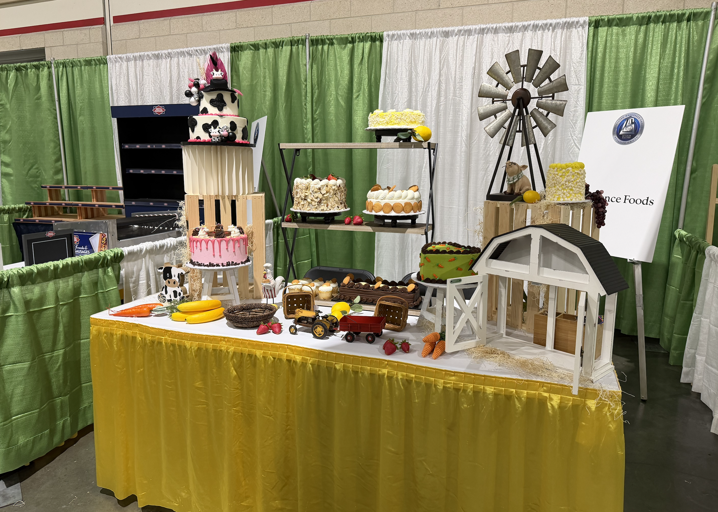 Cake table with various decorated cakes, toy farm animals, and farm-themed decorations, set against green and white curtains at an event.
