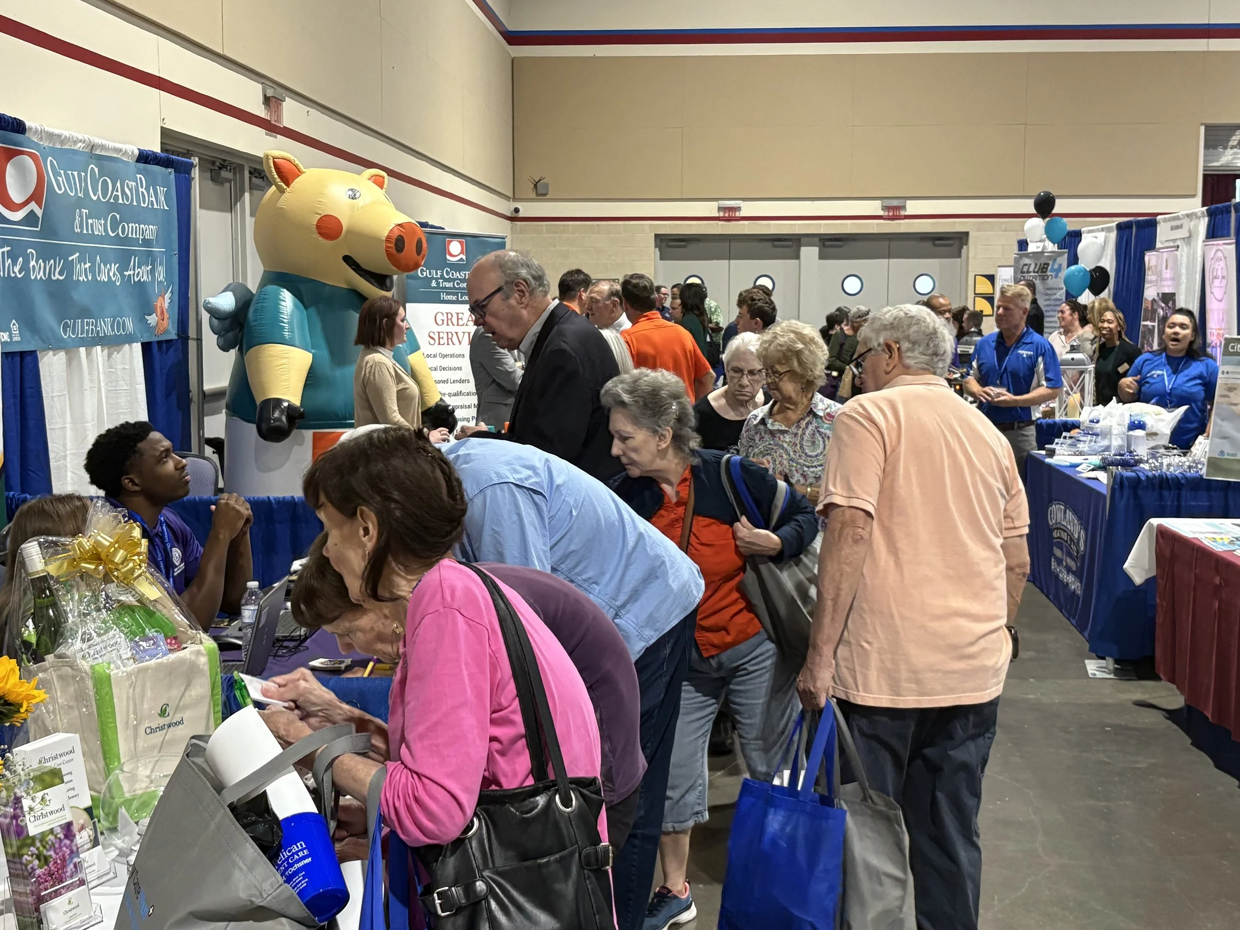 People browsing booths at an indoor expo or trade show, with a large inflatable pig mascot in the background, balloons, and promotional displays.