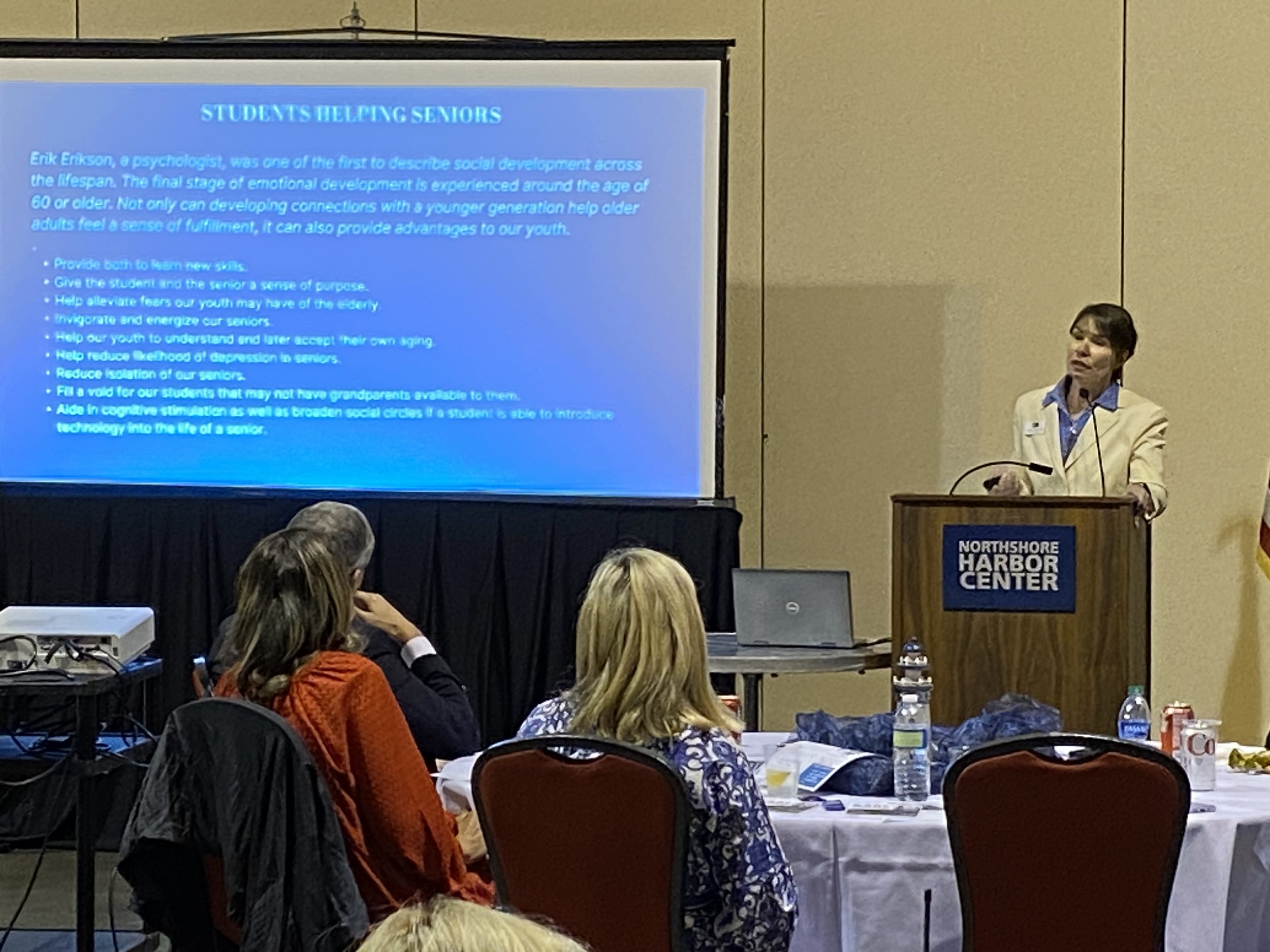 A woman standing at a podium delivering a presentation to an audience at Northshore Harbor Center. A large screen behind her displays a slide titled 'Students Helping Seniors' with bullet points about social development and aging.