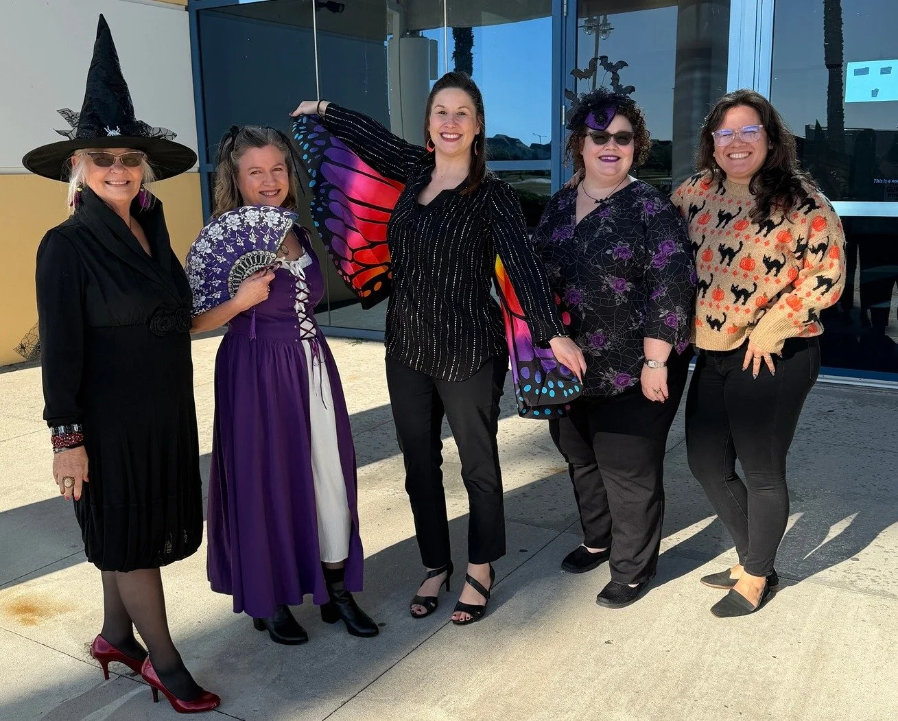 Six women dressed in colorful, whimsical attire standing outside in front of a modern glass building, with some wearing costumes and accessories like a witch hat, a fan, and butterfly wings, smiling at the camera.