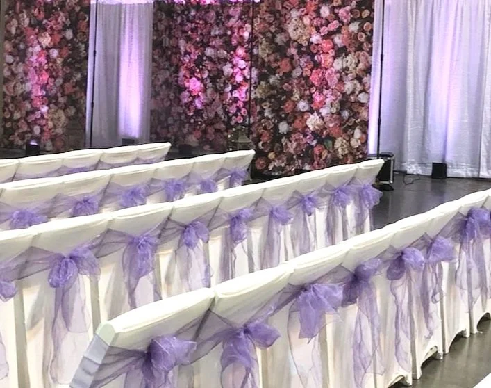 White chairs decorated with purple bows, facing a floral backdrop with pink, purple, and white flowers at an event or wedding venue.