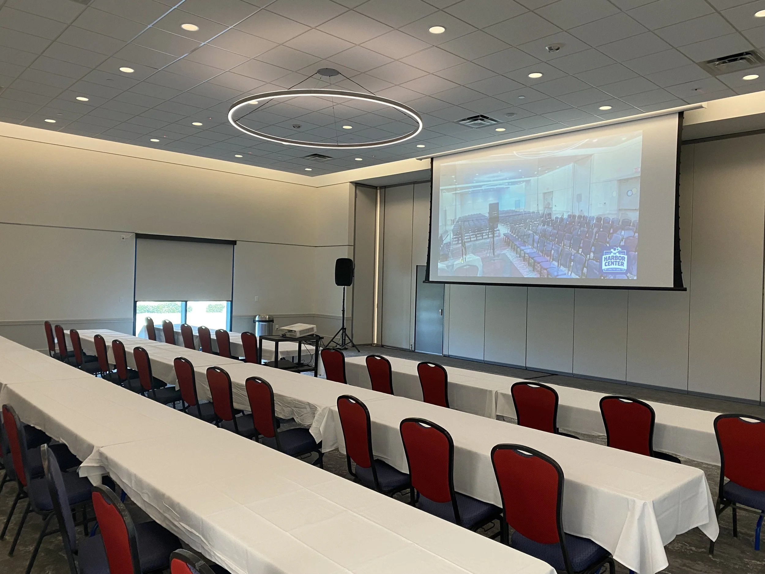 Conference room set up with red chairs around white covered tables, a large projection screen displaying an empty room with chairs and a logo that says Harbor Center, and a black speaker on a stand.
