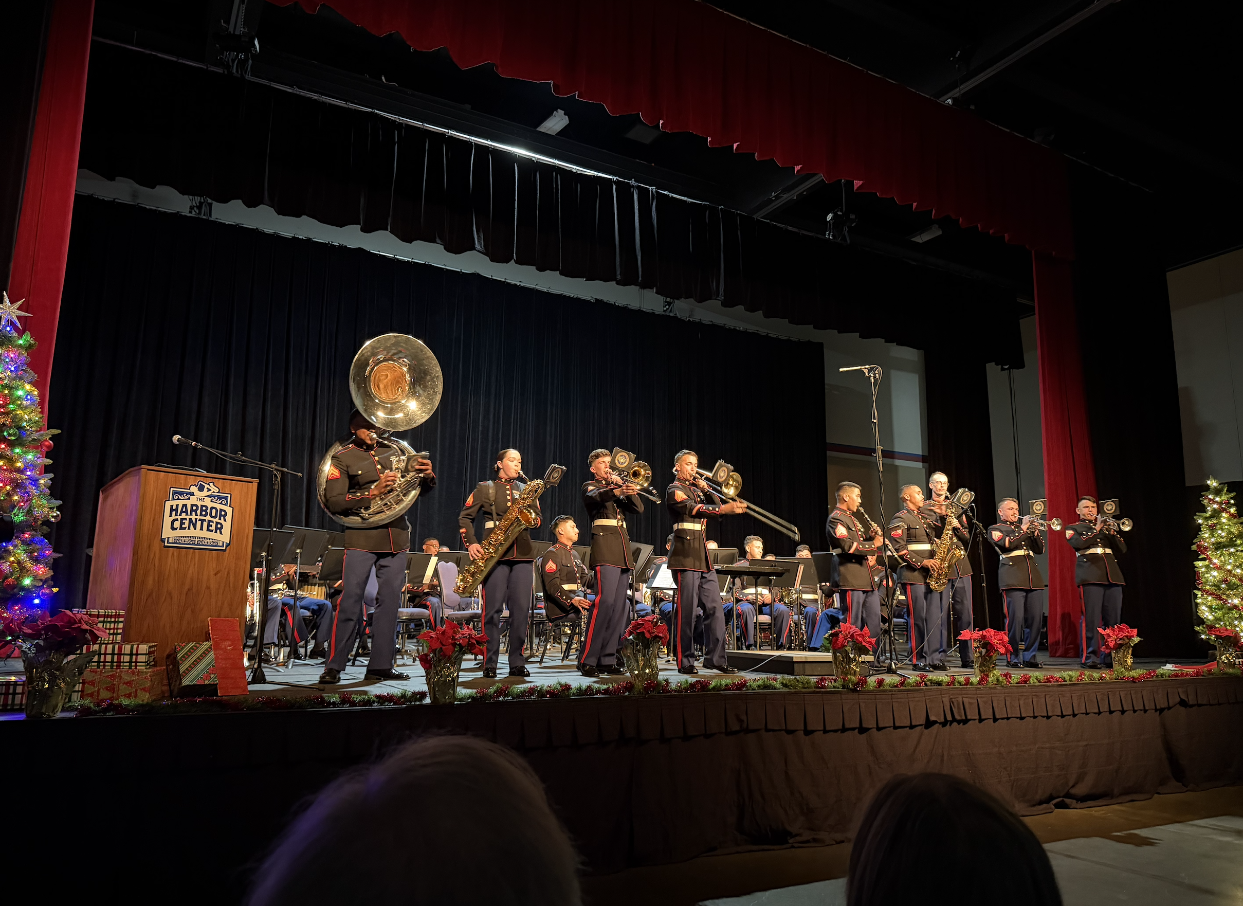 A military band performing on stage at the Harbor Center, decorated for Christmas with poinsettias and Christmas trees, during a holiday event.