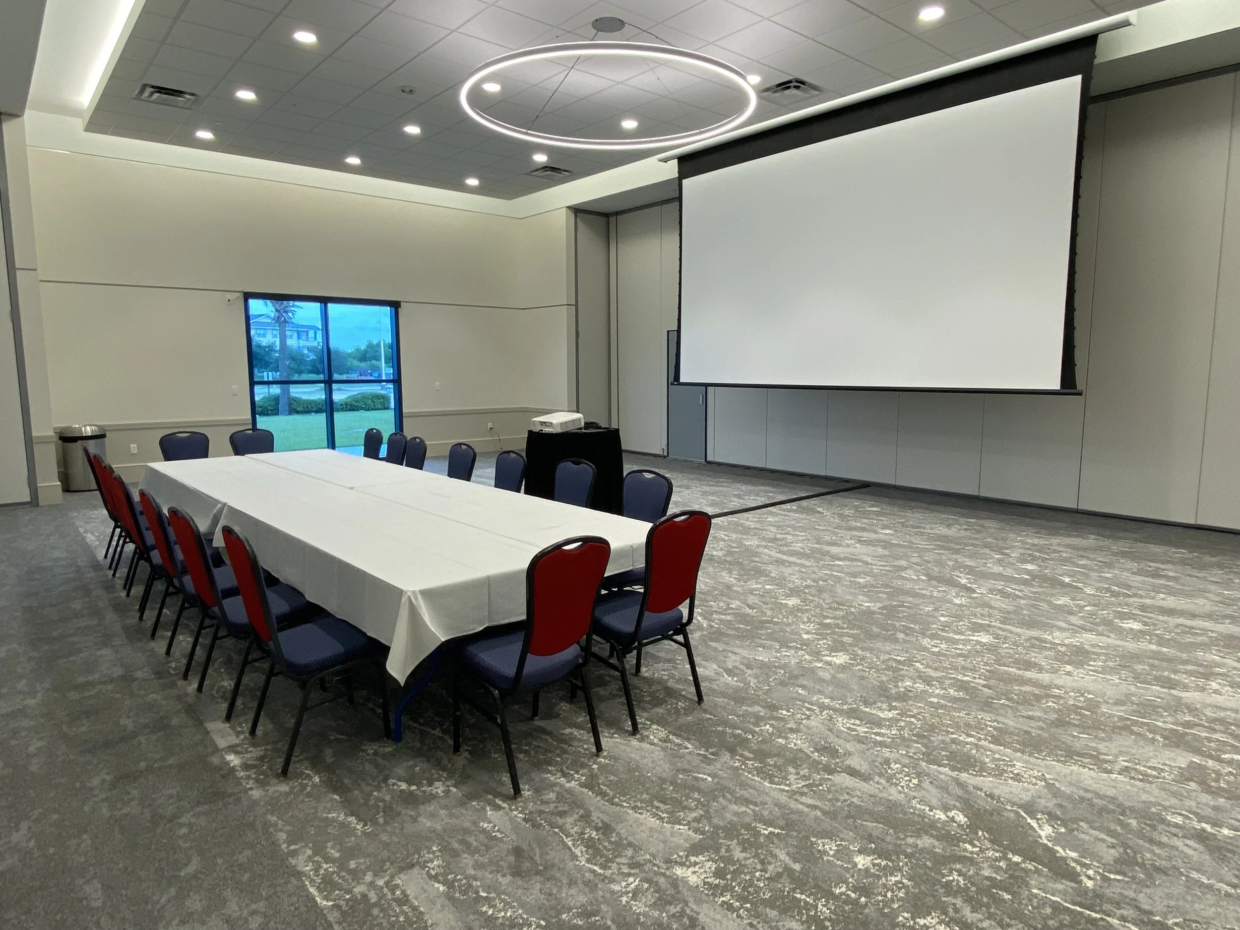 Empty conference room with a rectangular table covered with a white tablecloth, chairs around the table, a large projection screen on the wall, and a window showing an outdoor scene.