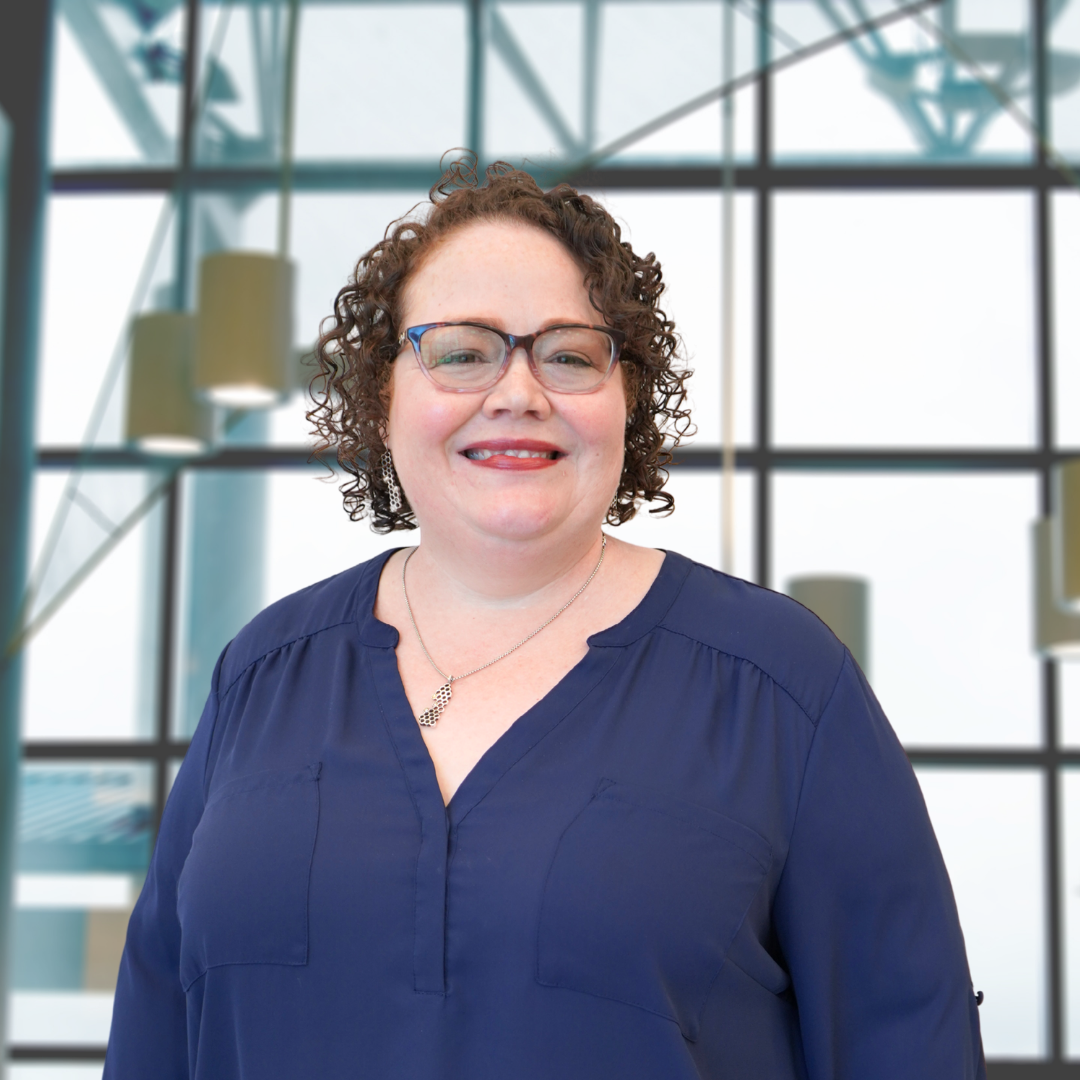 A smiling woman with curly brown hair and glasses, wearing a navy blue top, standing in front of a modern glass building.