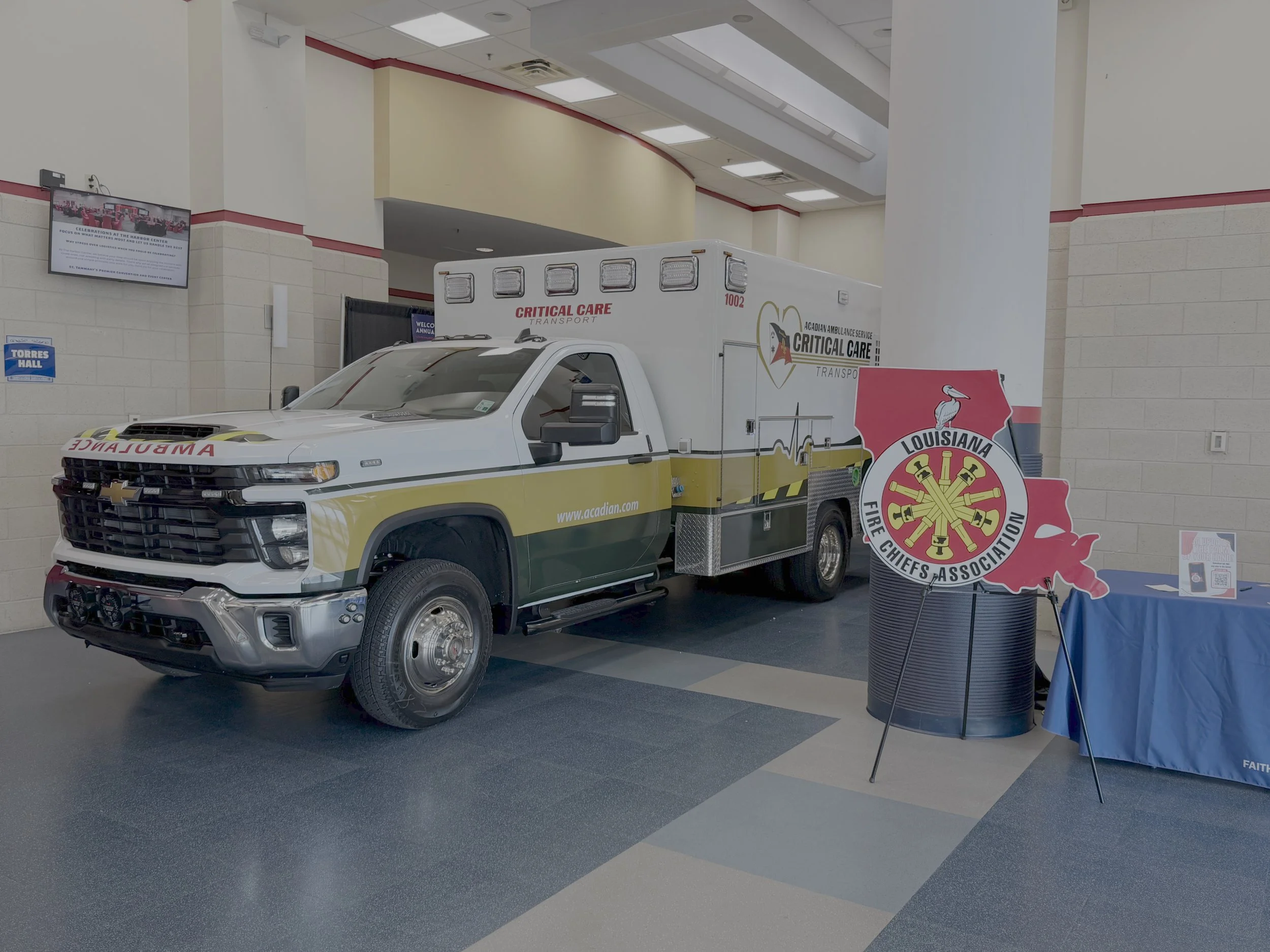 An emergency vehicle with 'Critical Care Transport' and 'Ambulance' markings, parked indoors at an event or exhibition space. Surrounding elements include a purple Louisiana Fire Chiefs & Association logo and various informational displays.