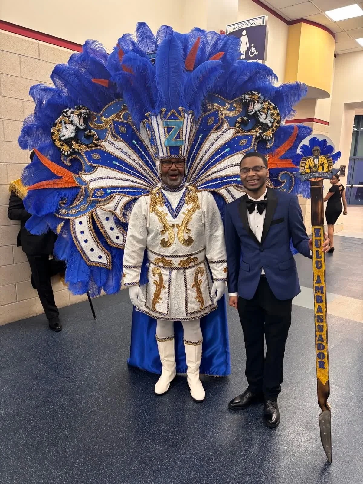 Two men in formal attire standing side by side; one is in an elaborate blue, gold, and white costume with feathers and a large decorative headdress, the other in a dark blue tuxedo with a bow tie. The man in costume holds a staff with a sign that say