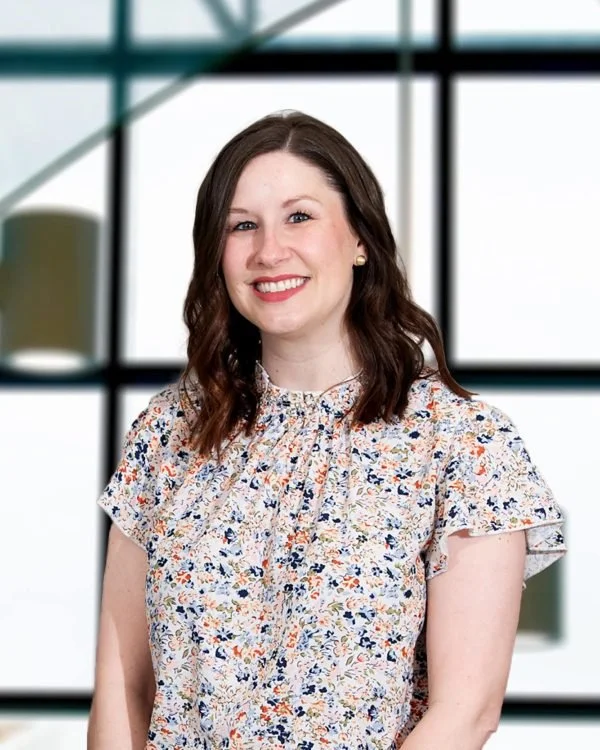 A woman with shoulder-length brown hair smiling, wearing a floral blouse, standing indoors with large windows and a blurred green plant in the background.
