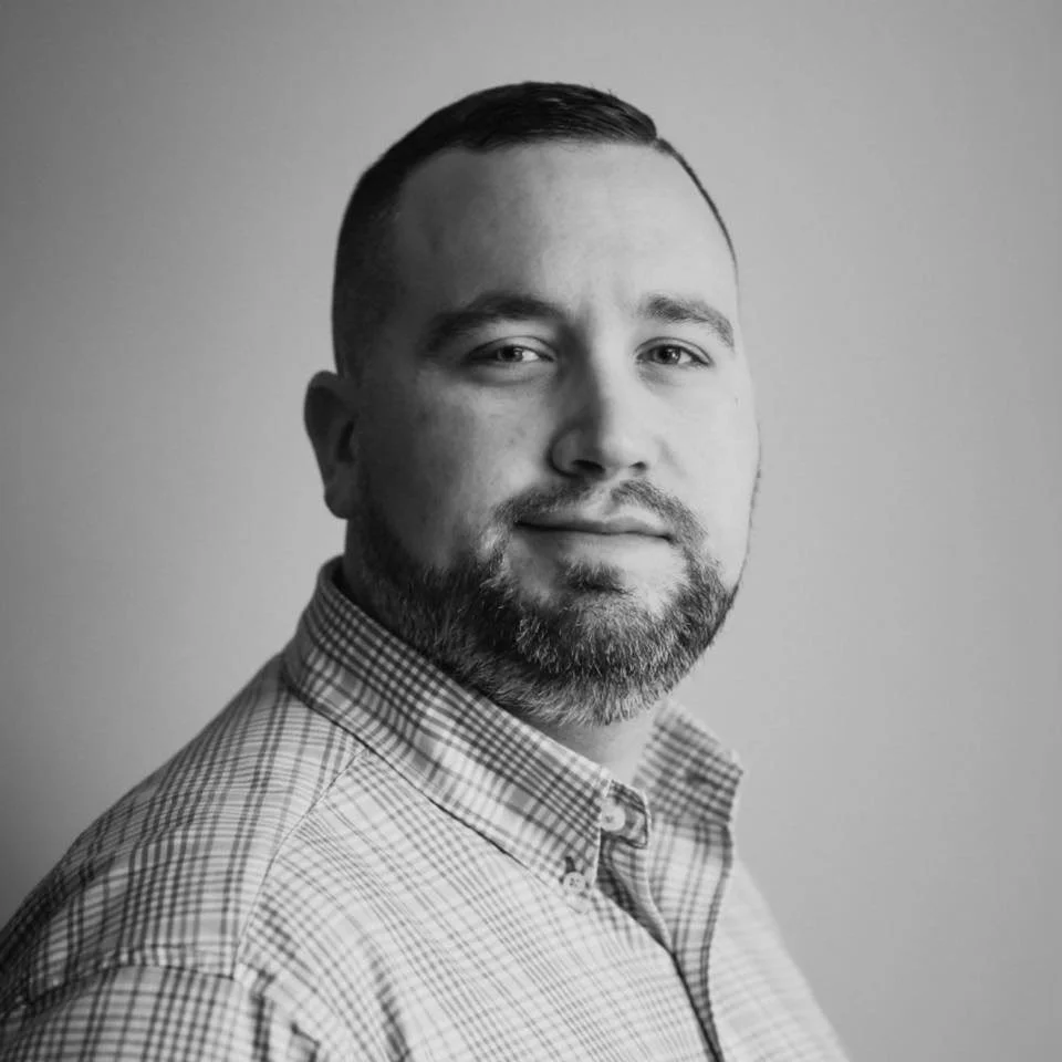 Black and white headshot of a man with short dark hair and a beard, wearing a checkered shirt, looking at the camera with a slight smile.