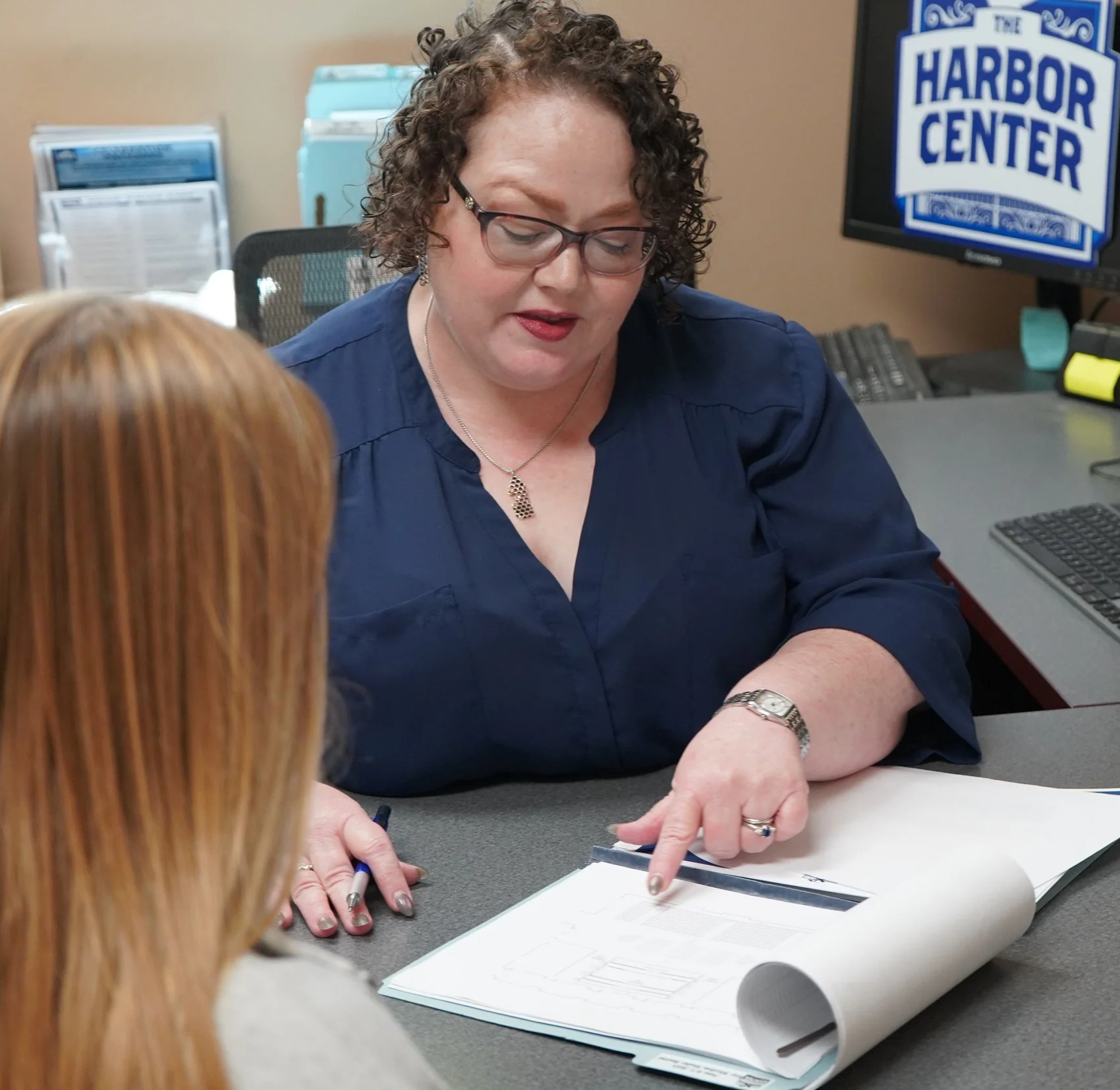 A woman with curly brown hair, glasses, and a navy blue blouse is sitting at a desk, pointing at a blueprint or document. There is a person with red hair sitting across from her. In the background, there is a monitor displaying the logo of The Harbor Center.