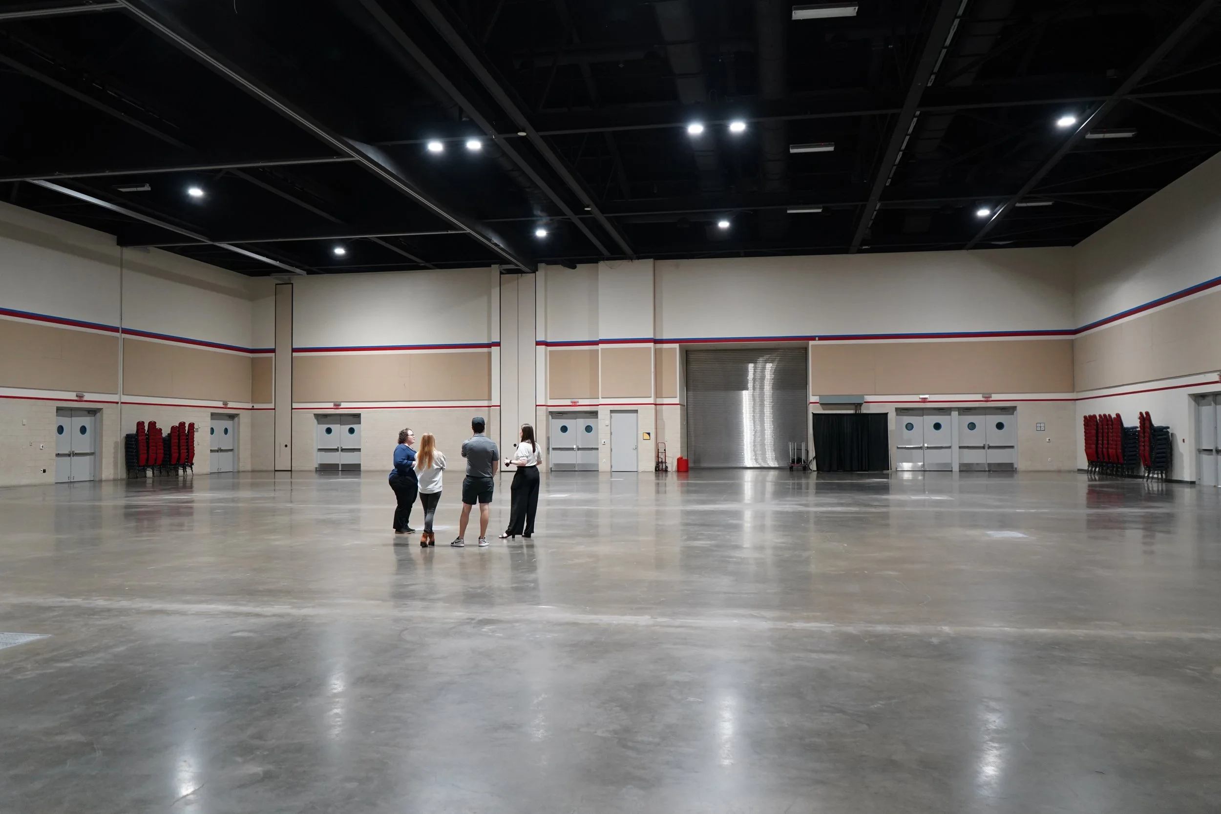 Four people stand and talk in the middle of a large, empty indoor event space with a polished concrete floor, beige walls decorated with red, white, and blue stripes, stacks of chairs against the walls, and a large metal door at the back.