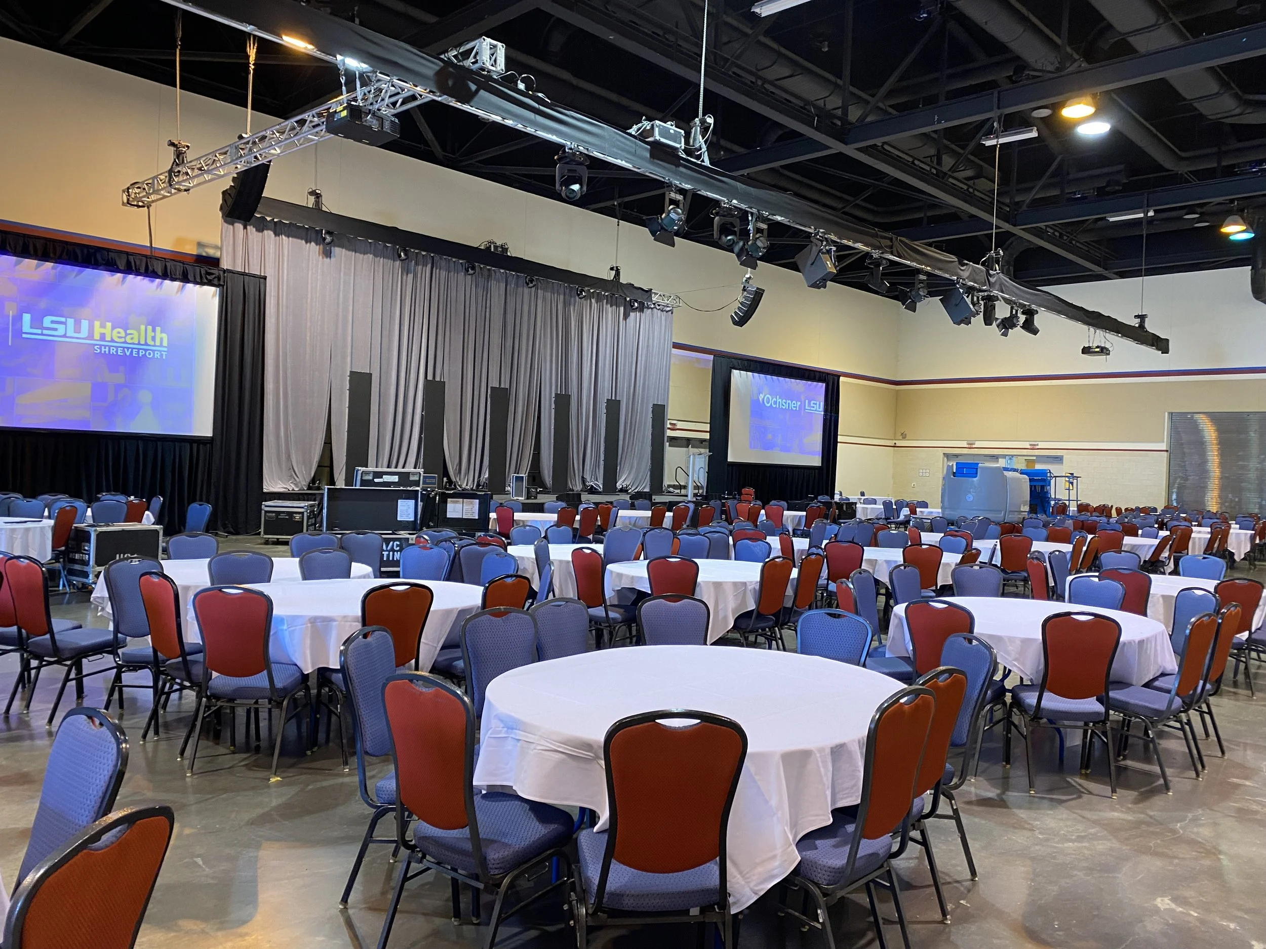 Conference room set up with round tables covered with white tablecloths, arranged in front of a stage with curtains and large screens displaying LSU Health Shreveport logos.