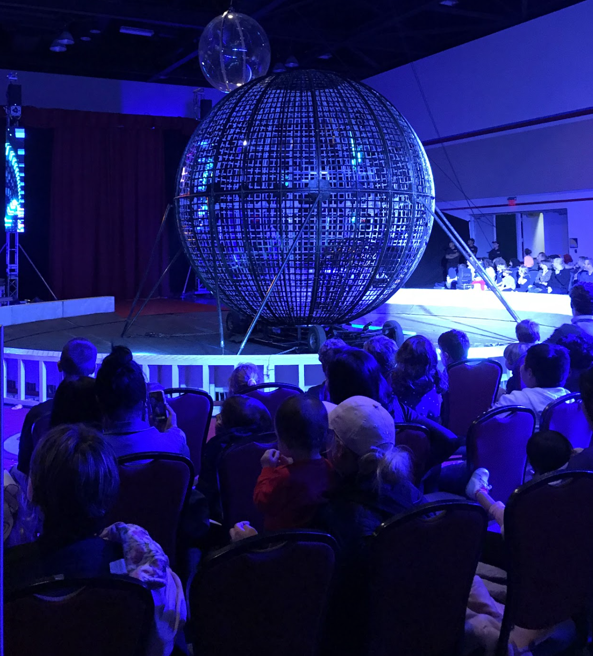 A large sphere made of metal grid resembling a globe on a stage, surrounded by people watching a performance or presentation in a dimly lit room with blue lighting.