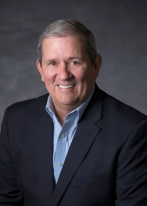 Smiling middle-aged man in a dark suit and light blue shirt against a dark gray background.