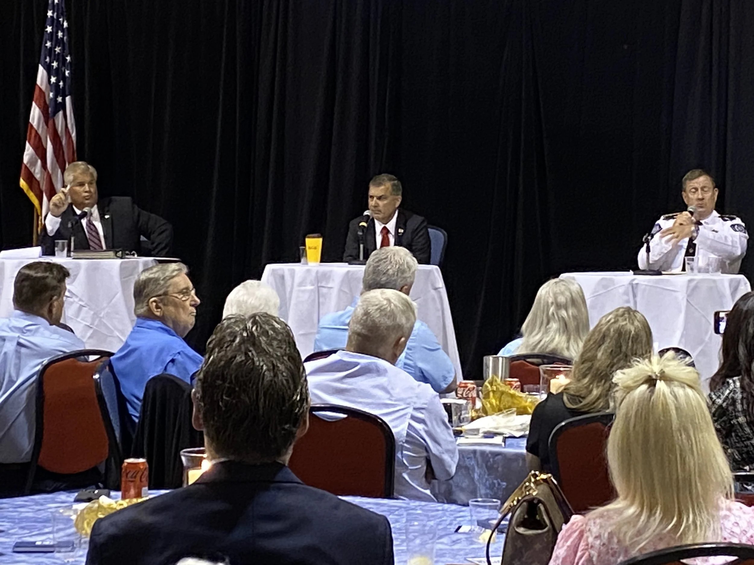 Panel discussion with three men on stage, two at tables with microphones, audience seated at tables in front, American flag in background.