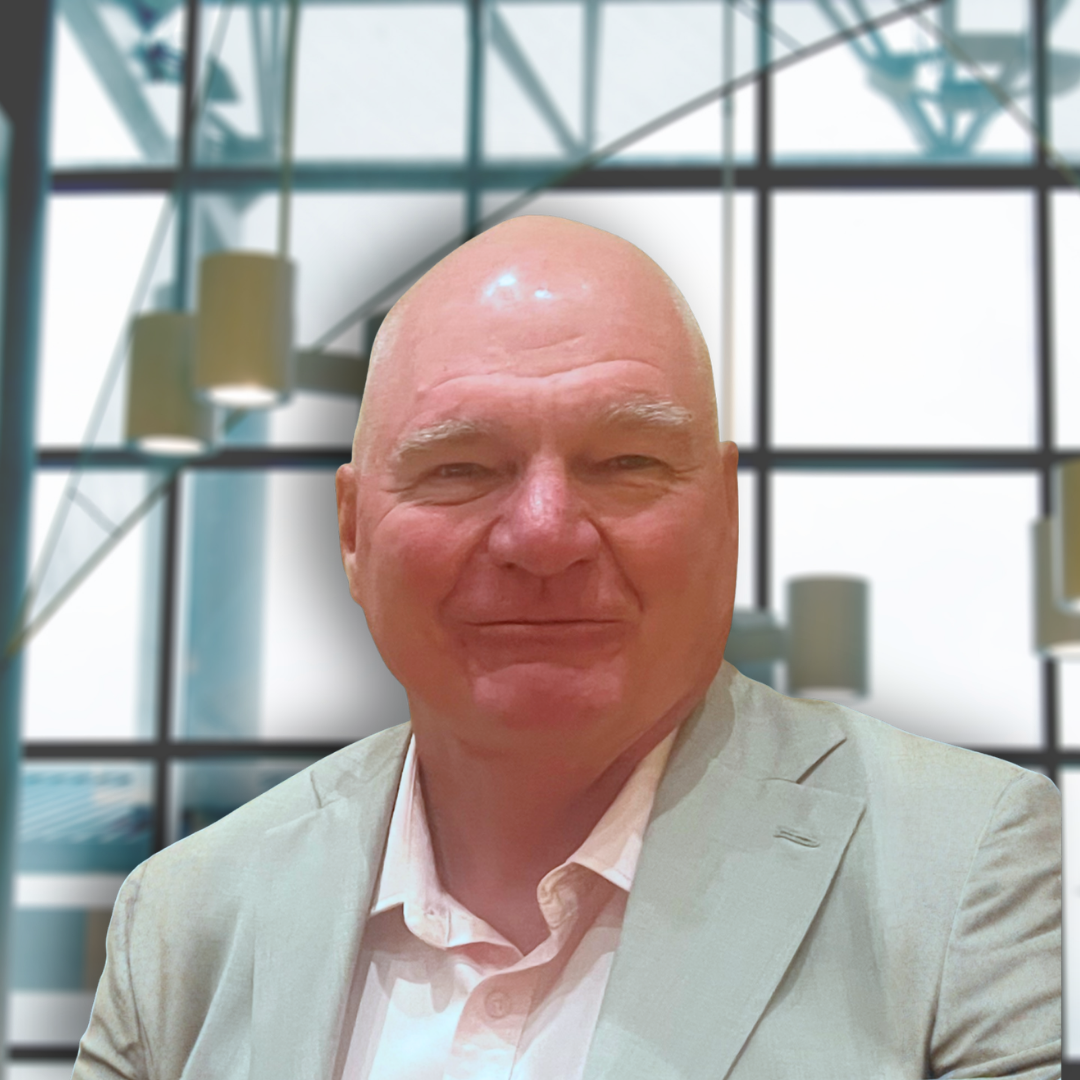 A man in a light-colored suit and white shirt smiling in front of a glass window in a modern building.