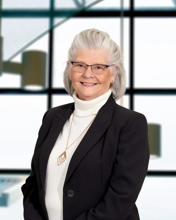 A smiling elderly woman with gray hair and glasses, wearing a black blazer and white turtleneck, standing in front of a large window with a grid pattern.