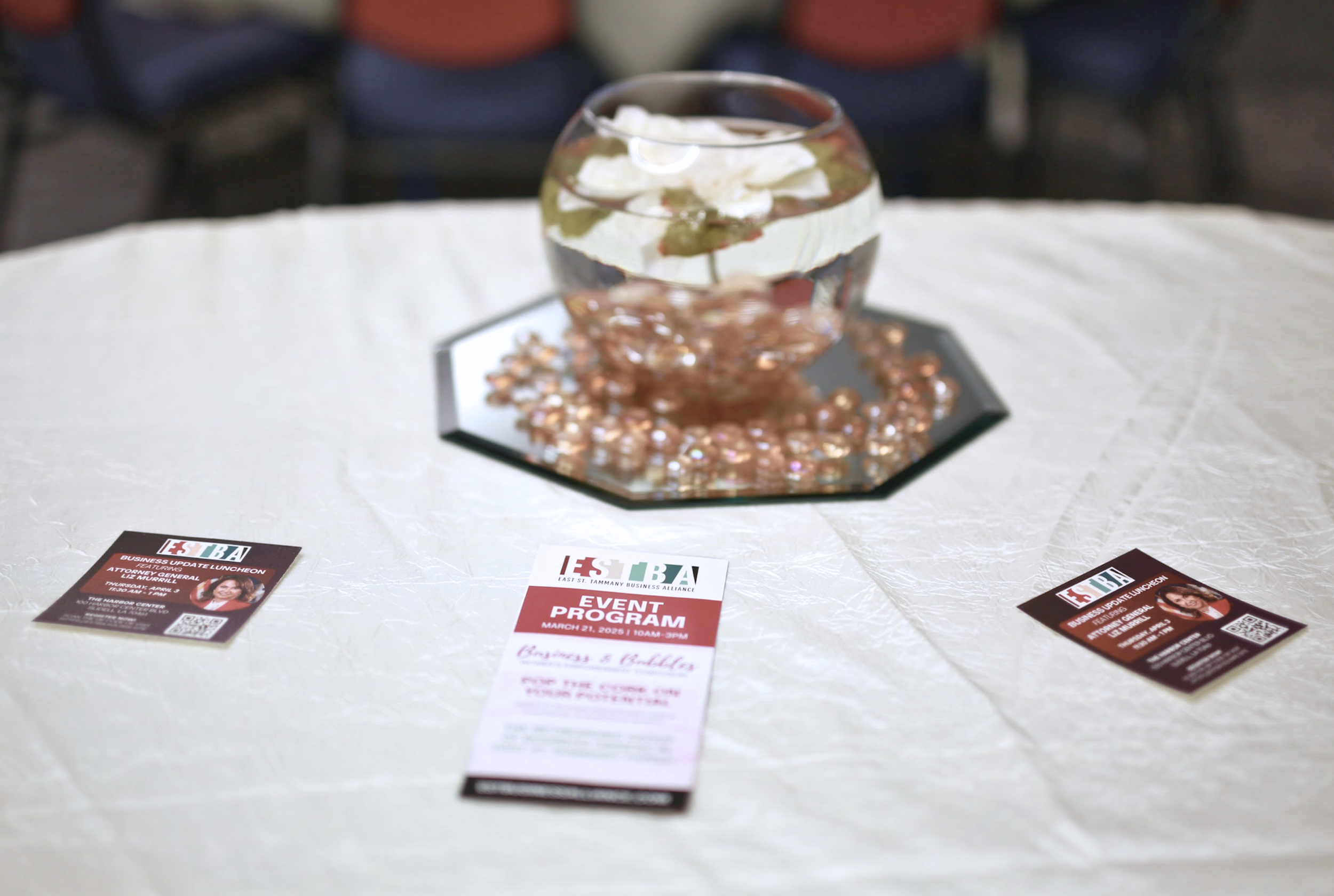 A glass of water with a flower inside, placed on a reflective octagonal mirror, on a table covered with a white tablecloth, with event flyers nearby.
