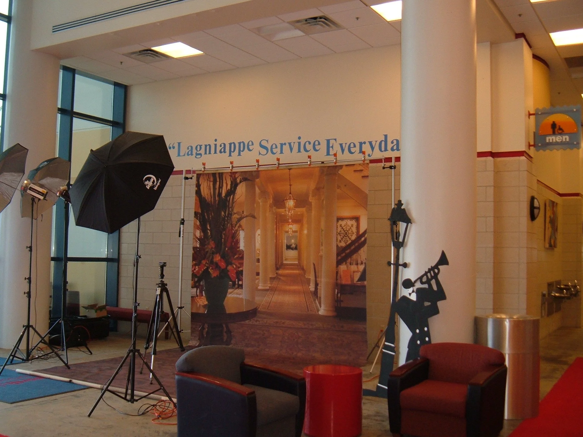 Indoor scene of photography setup with umbrellas, tripods, and cutout figures, near a large banner with a hallway image and text that says 'Lagniappe Service Everyda' in a public building or hotel lobby.
