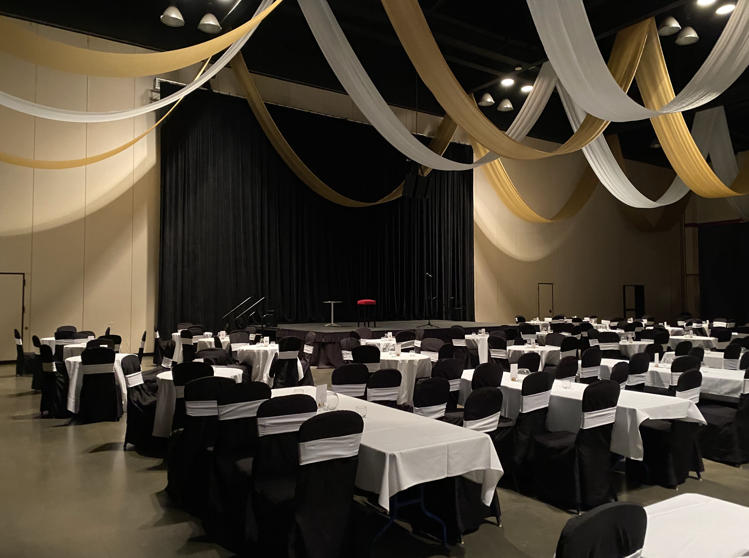 Banquet hall with round tables covered with white tablecloths, black chairs with white sashes, a stage with black curtains, and flowing fabric decorations in gold and white hanging from the ceiling.