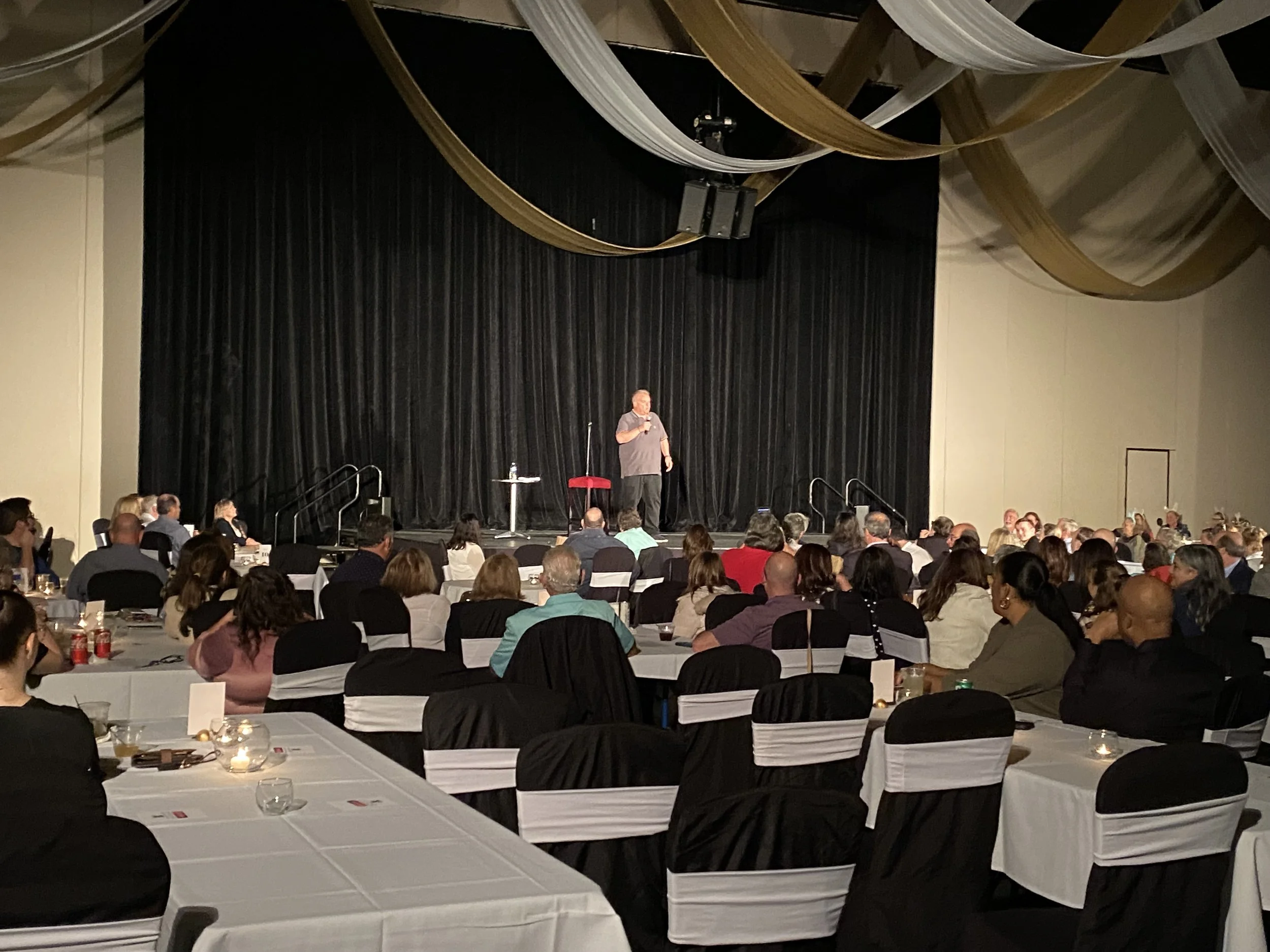 A man speaking on stage at a conference or event, with an audience seated at round tables in a banquet hall. The stage has black curtains and is decorated with large gold and white drapes hanging from the ceiling.