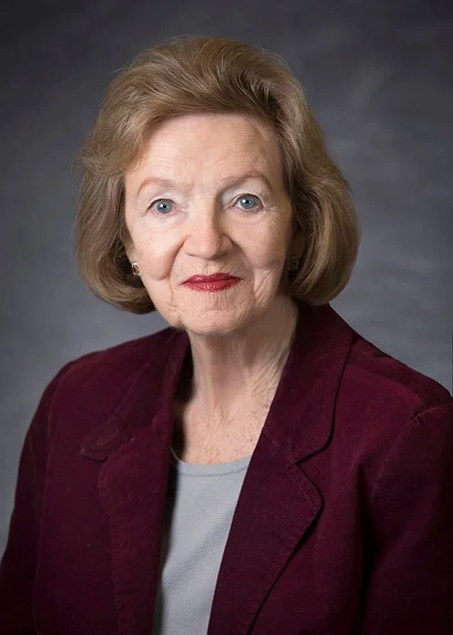 A portrait of an elderly woman with light skin, short wavy blonde hair, blue eyes, wearing a burgundy blazer and a light gray top, against a dark gray background.