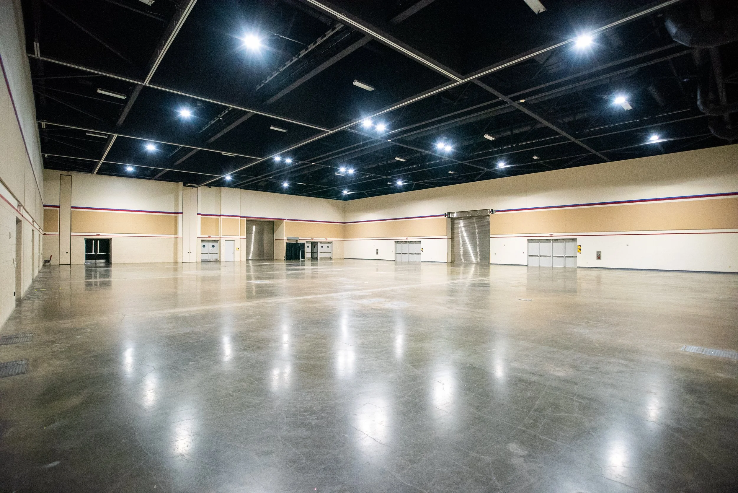 Empty large indoor space with polished concrete floor, beige walls with red and blue trim, and a black ceiling with multiple bright white lights.