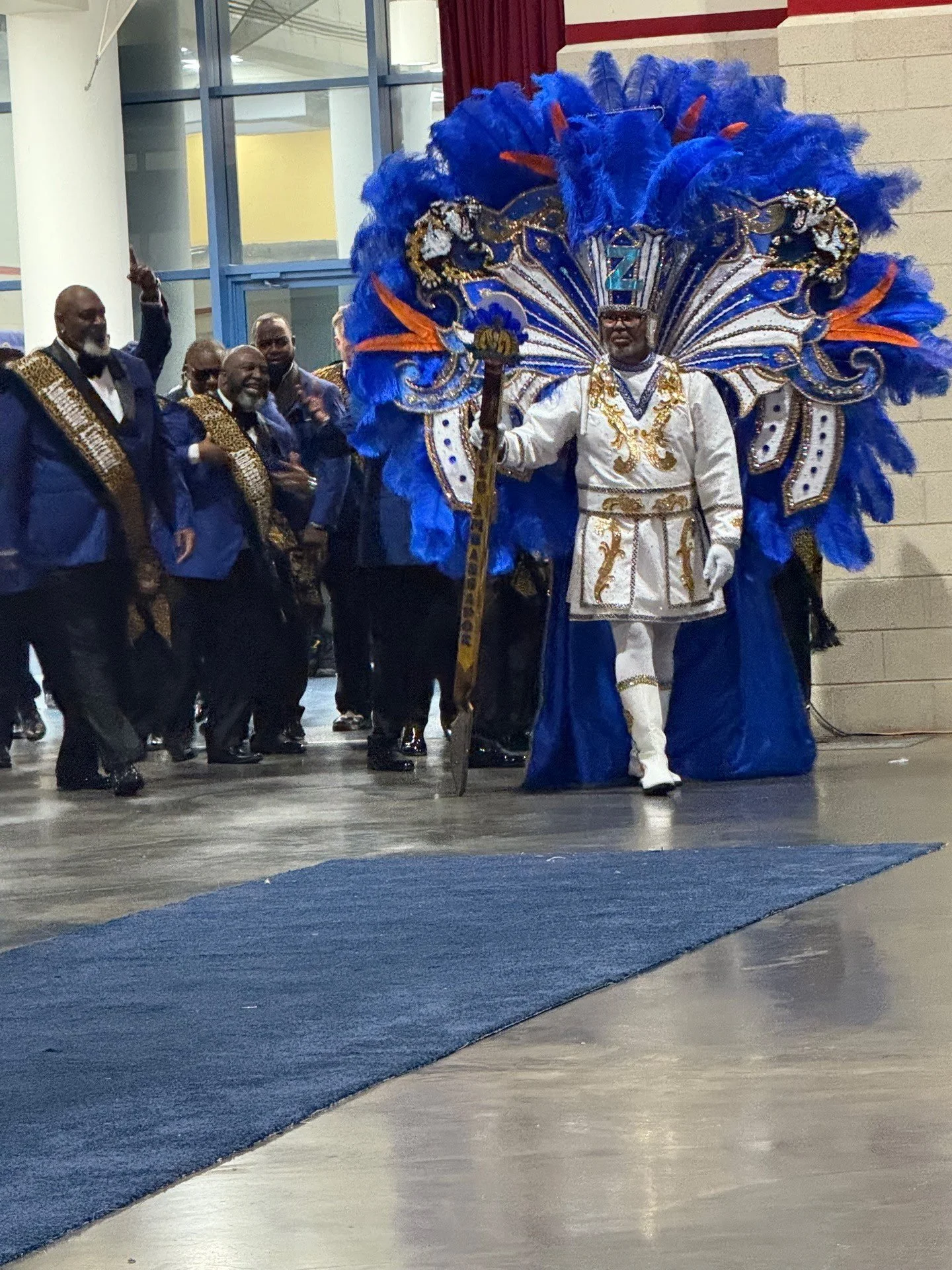 A man dressed in an elaborate costume with blue feathers and gold accents, holding a staff, leading a procession of men in navy blue suits with gold sashes.