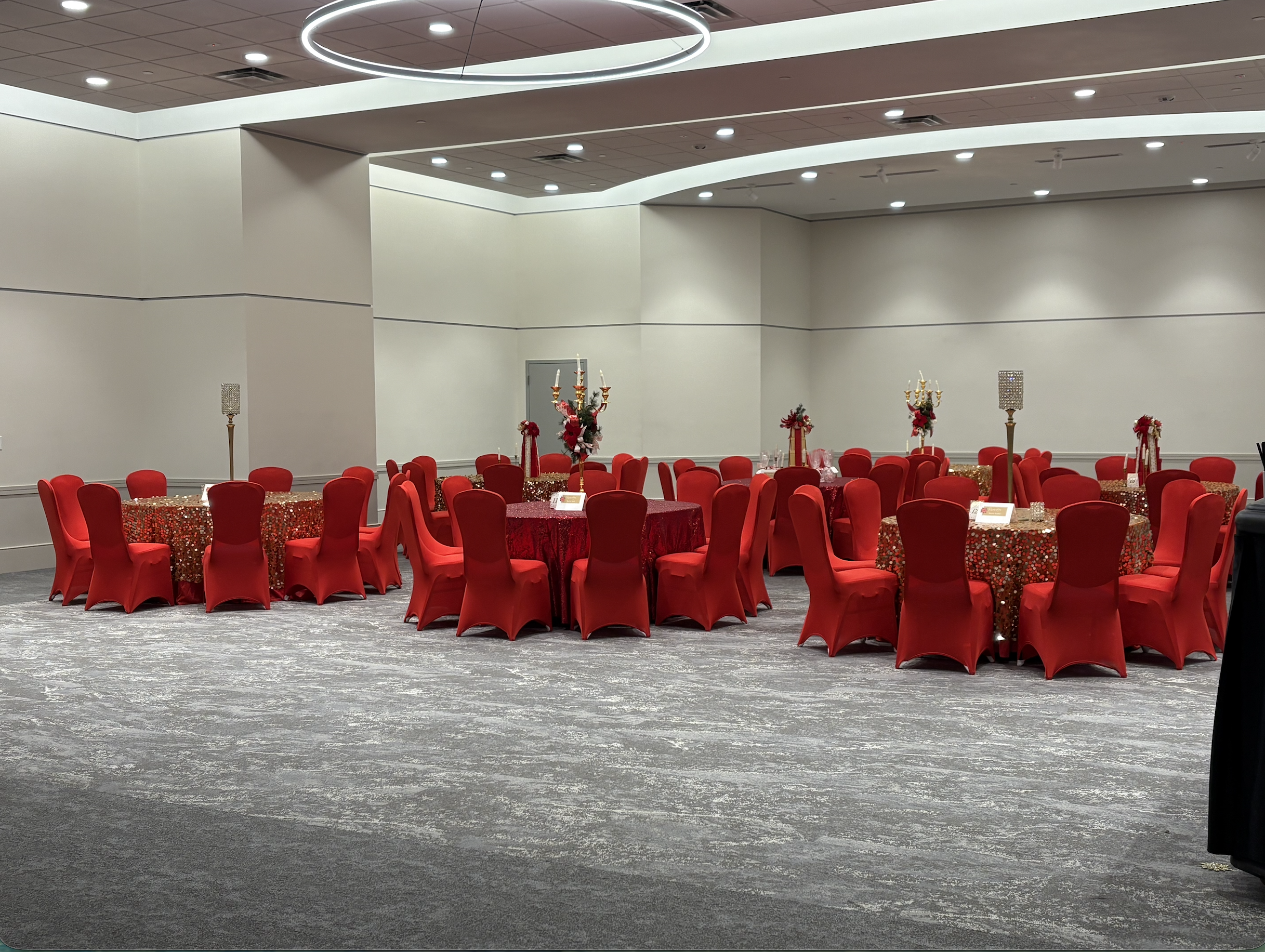 Event hall decorated with round tables covered in red tablecloths, surrounded by red chairs, with gold and red centerpieces and candelabras, ready for a celebration or gathering.