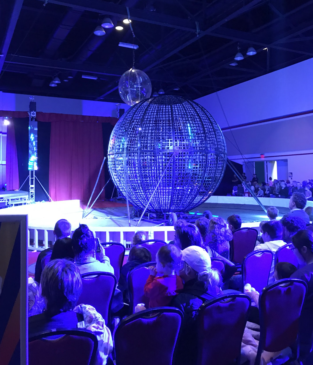 Audience watching a circus act featuring a large metallic globe on stage with blue lighting