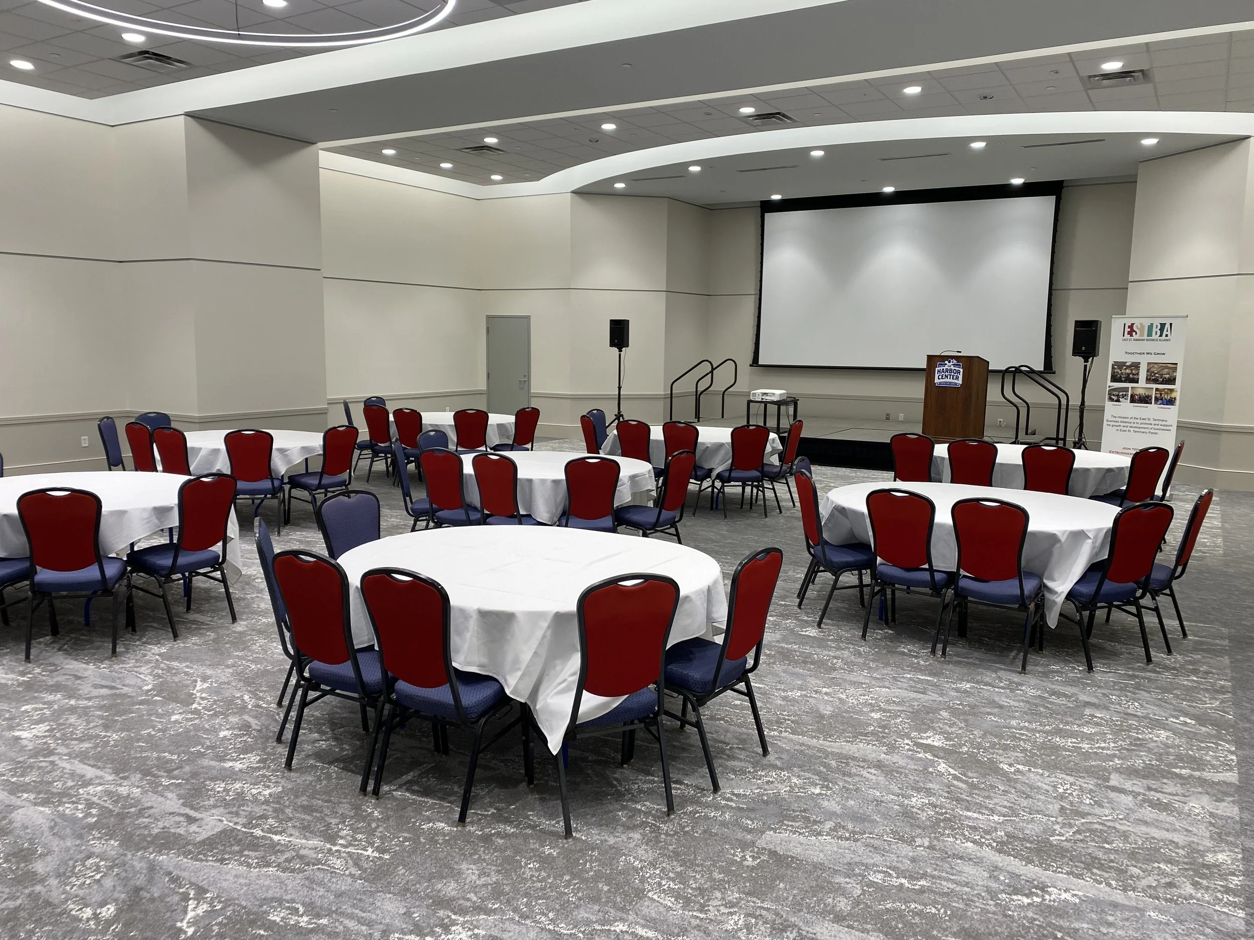 Event space with round tables covered in white tablecloths and red and navy chairs, a stage with a large projection screen, speakers, and a podium, set up for a conference or event.