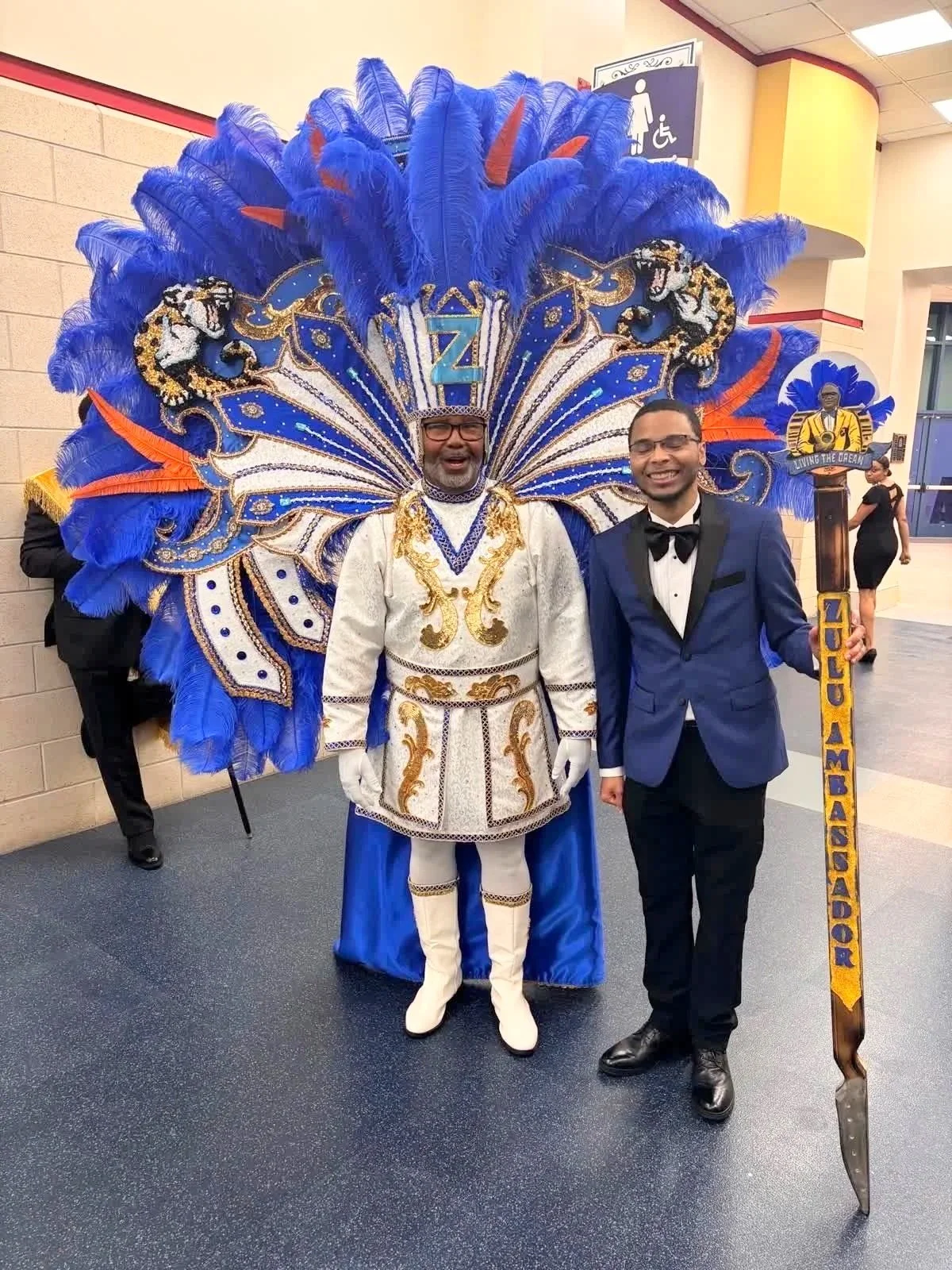 Two men are dressed in formal and colorful costumes at an event, one wearing a large, elaborate blue and gold feathered headdress and traditional attire, the other in a blue tuxedo with a bow tie, holding a staff that reads 'HOU AMAHANOR'.