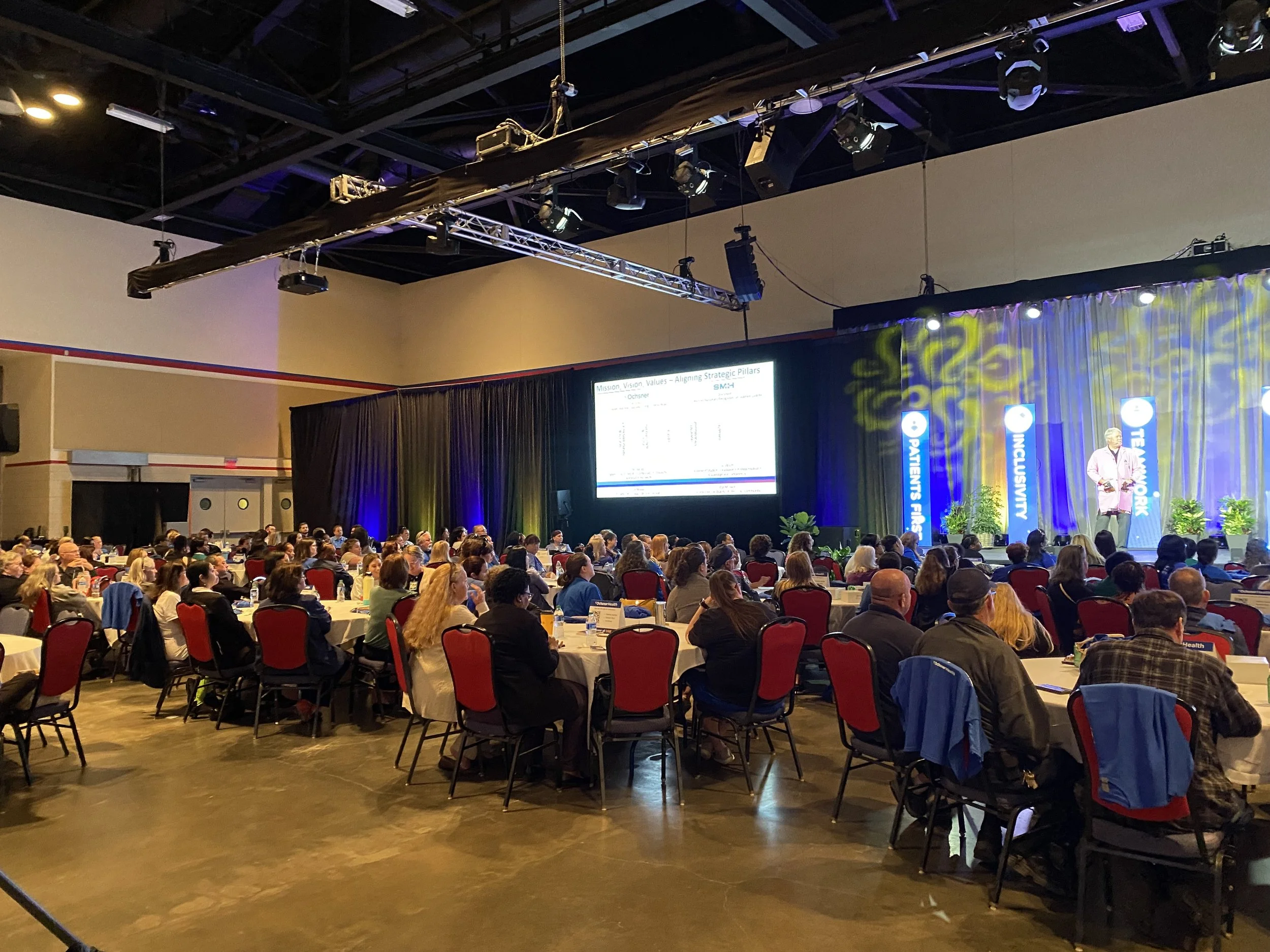 Large conference room filled with attendees seated at round tables, facing a stage with a speaker and a presentation screen, during a professional event.