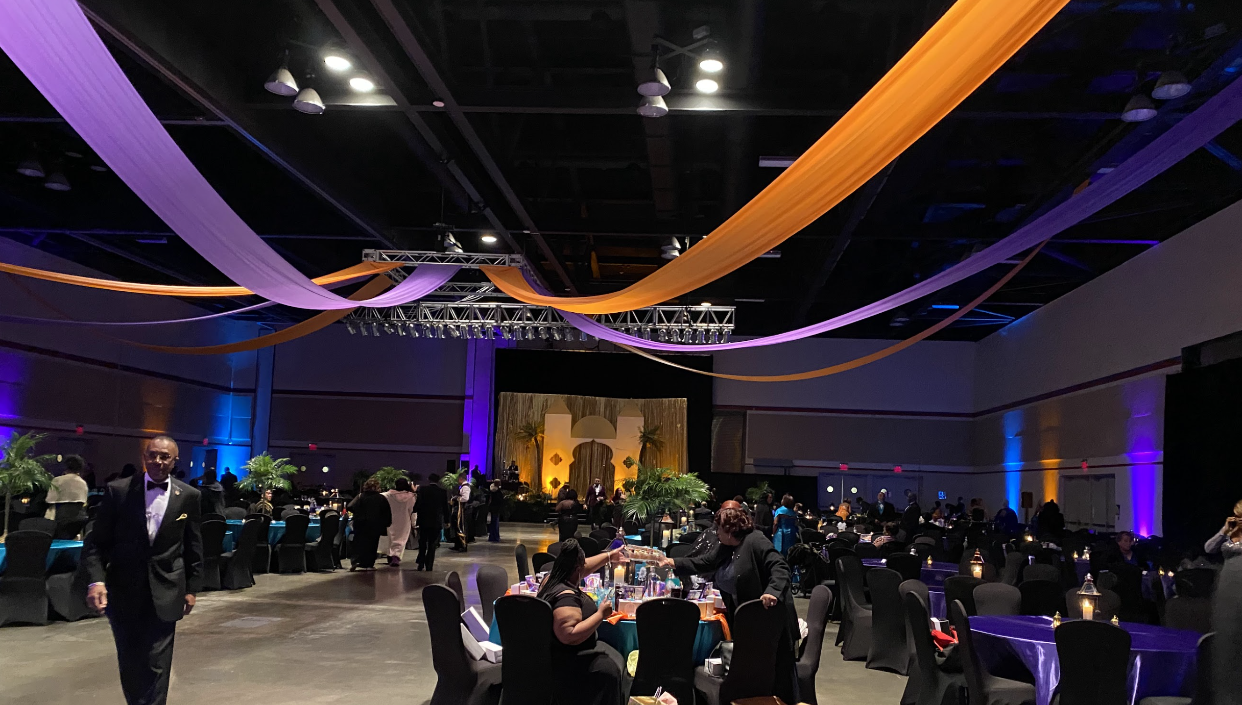 A decorated banquet hall with purple and orange drapes hanging from the ceiling, a stage with gold curtains, and tables set with candles and floral centerpieces, filled with guests dressed in formal attire.